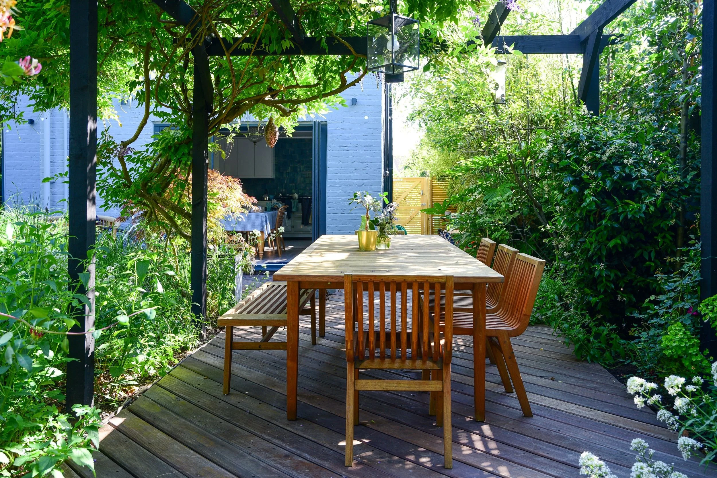 A wooden dining table and chairs sit beneath a black timber pergola draped in wisteria and climbing plants in a Teddington garden, with a glimpse of the blue-painted kitchen extension beyond. JaneShanksterGardenDesign.co.uk.

Dappled light filters th