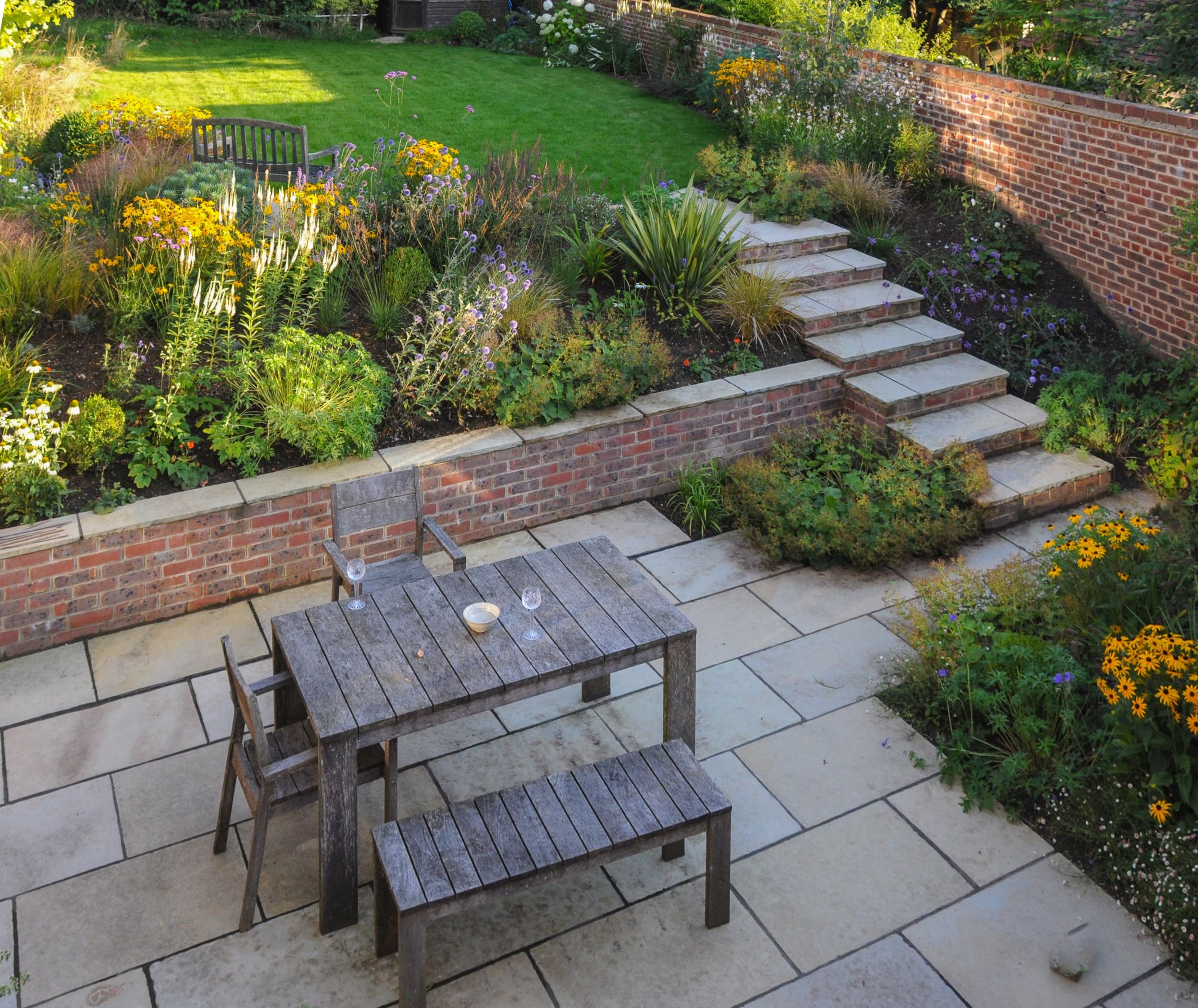 An elevated view shows a dining terrace, brick steps and sweeping lawn bordered by colourful pollinator planting. JaneShanksterGardenDesign.co.uk

Seen from above, the garden's layout unfolds: a stone terrace set for dining, brick steps rising throug