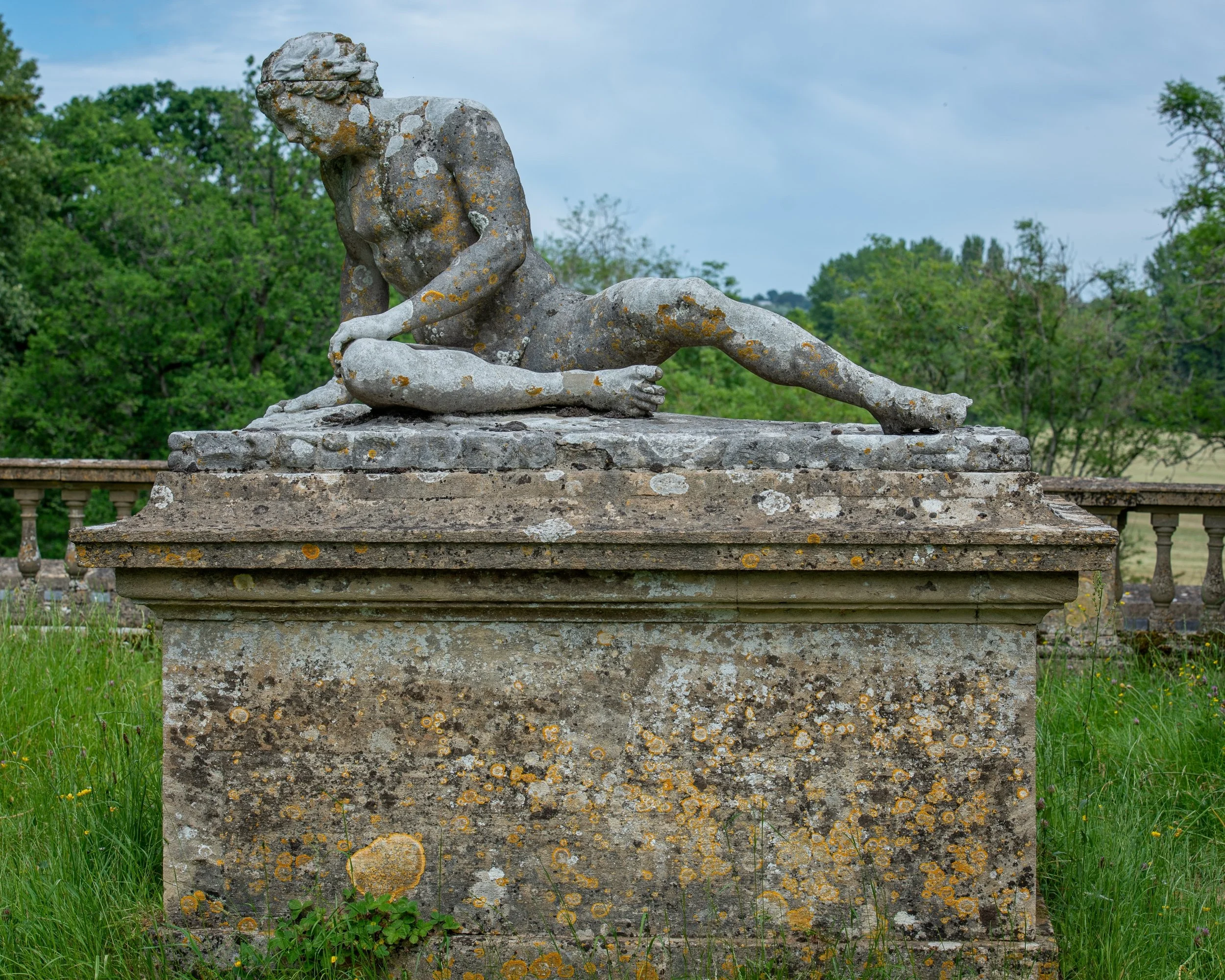 Lichen-covered statue of the Dying Gladiator on a stone plinth at Rousham, with a balustrade and trees behind.