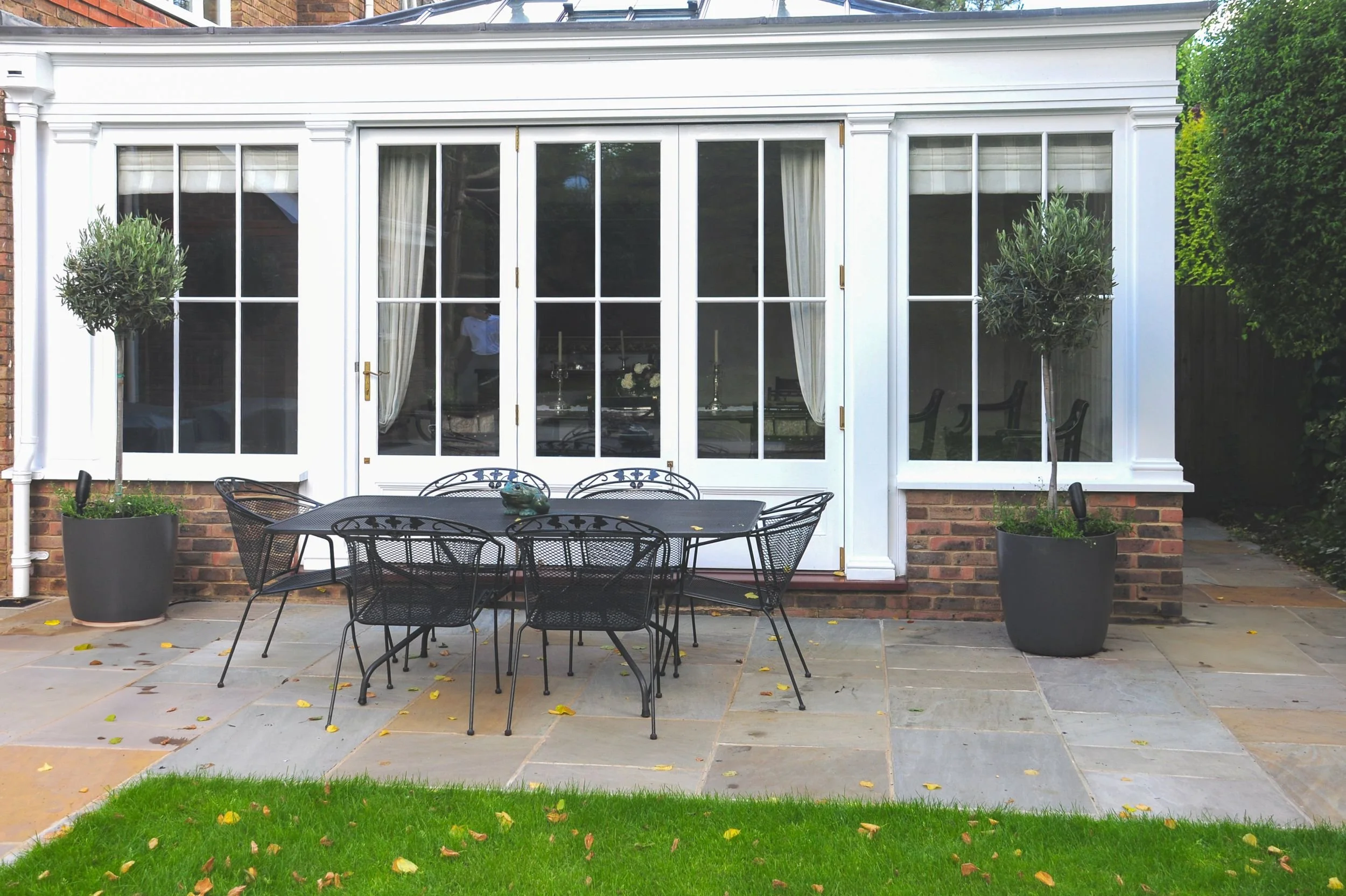 A wrought-iron dining table sits on a stone terrace outside the orangery, flanked by potted olive trees. JaneShanksterGardenDesign.co.uk

An elegant wrought-iron dining set is arranged on a stone terrace beneath the orangery's glazed doors, with clip