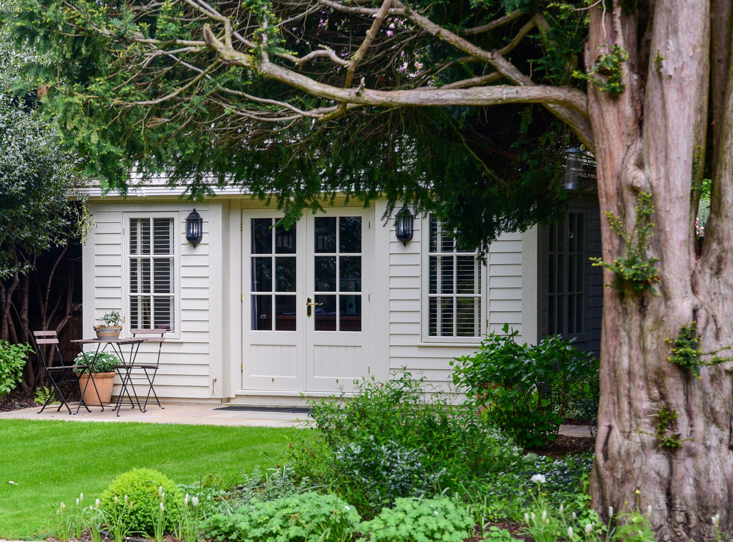 The Crane Garden Building is glimpsed through mature yew trees beyond a terrace bistro set. JaneShanksterGardenDesign.co.uk

A glimpse through dark yew branches reveals the cream weatherboarded Crane Garden Building, with a small bistro table set on 