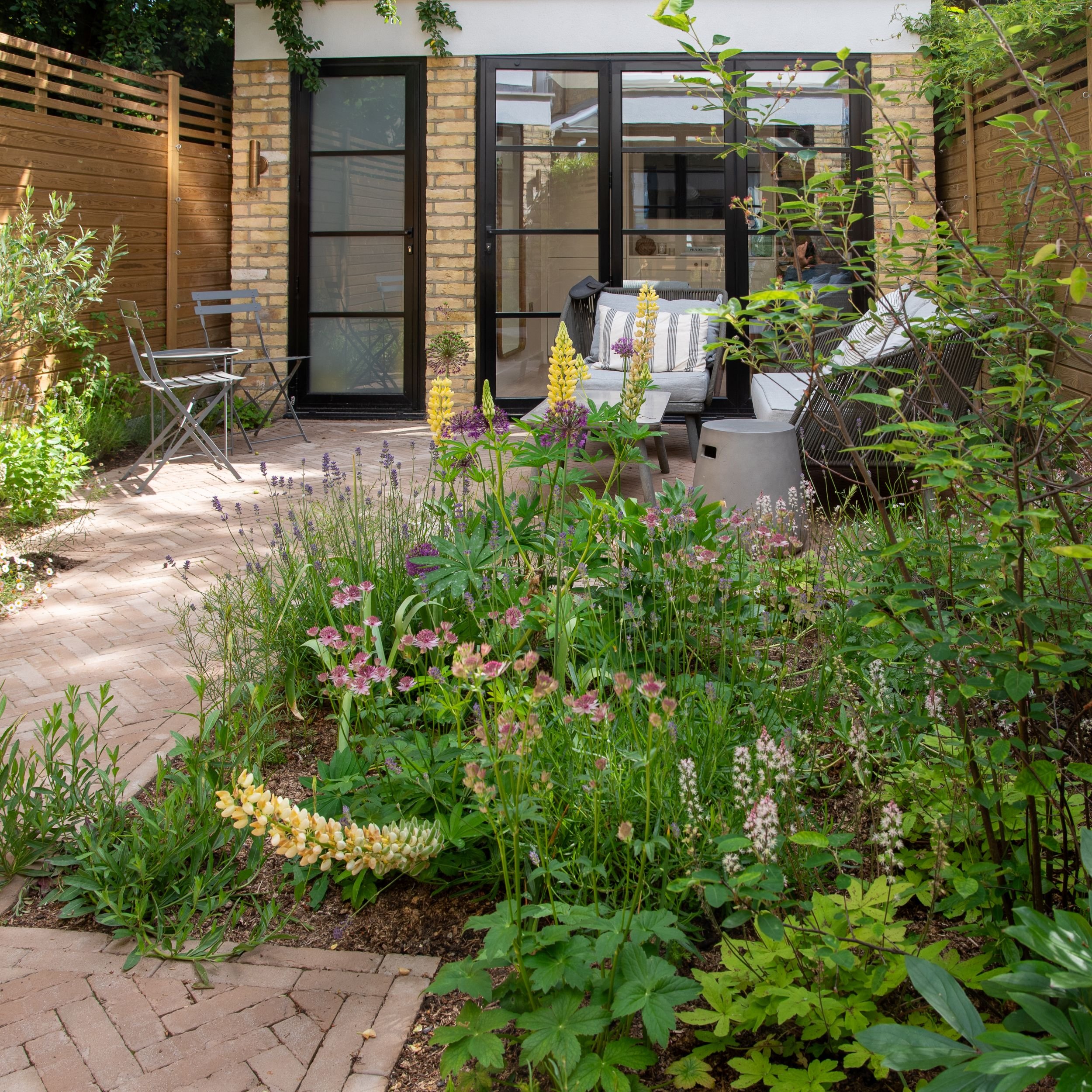 A full overview of the Fulham courtyard garden in summer, with herringbone brick paving, naturalistic planting borders of lupins, astrantia and thalictrum, bistro table and wicker sofa seating areas, cedar fencing and Crittall-style glazed doors to t