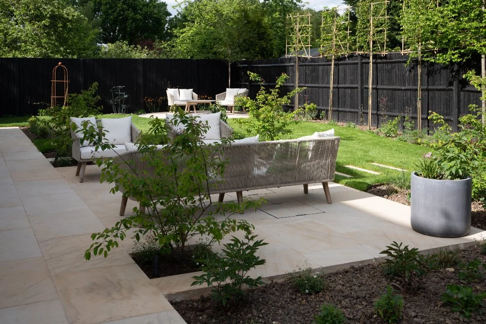Newly completed garden immediately after planting, with young trees, bare borders and rattan seating areas on a stone terrace.