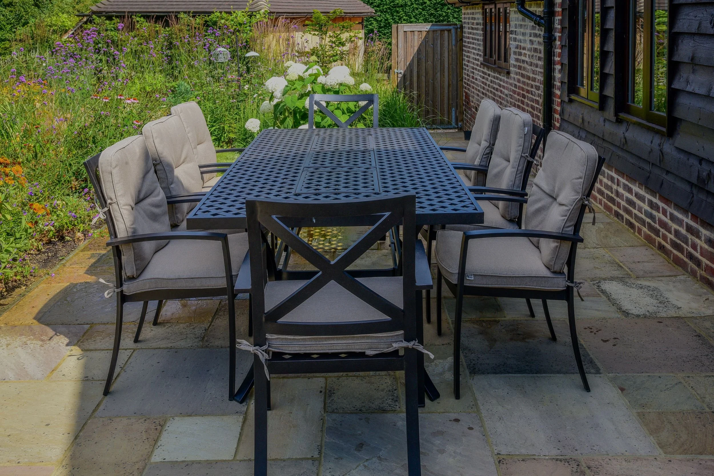 A black metal dining table and chairs sit on a stone patio beside a border of verbena and hydrangea in a Surrey garden. JaneShanksterGardenDesign.co.uk.

Tucked beside the house, this dining area sits close enough to the borders that verbena and hydr