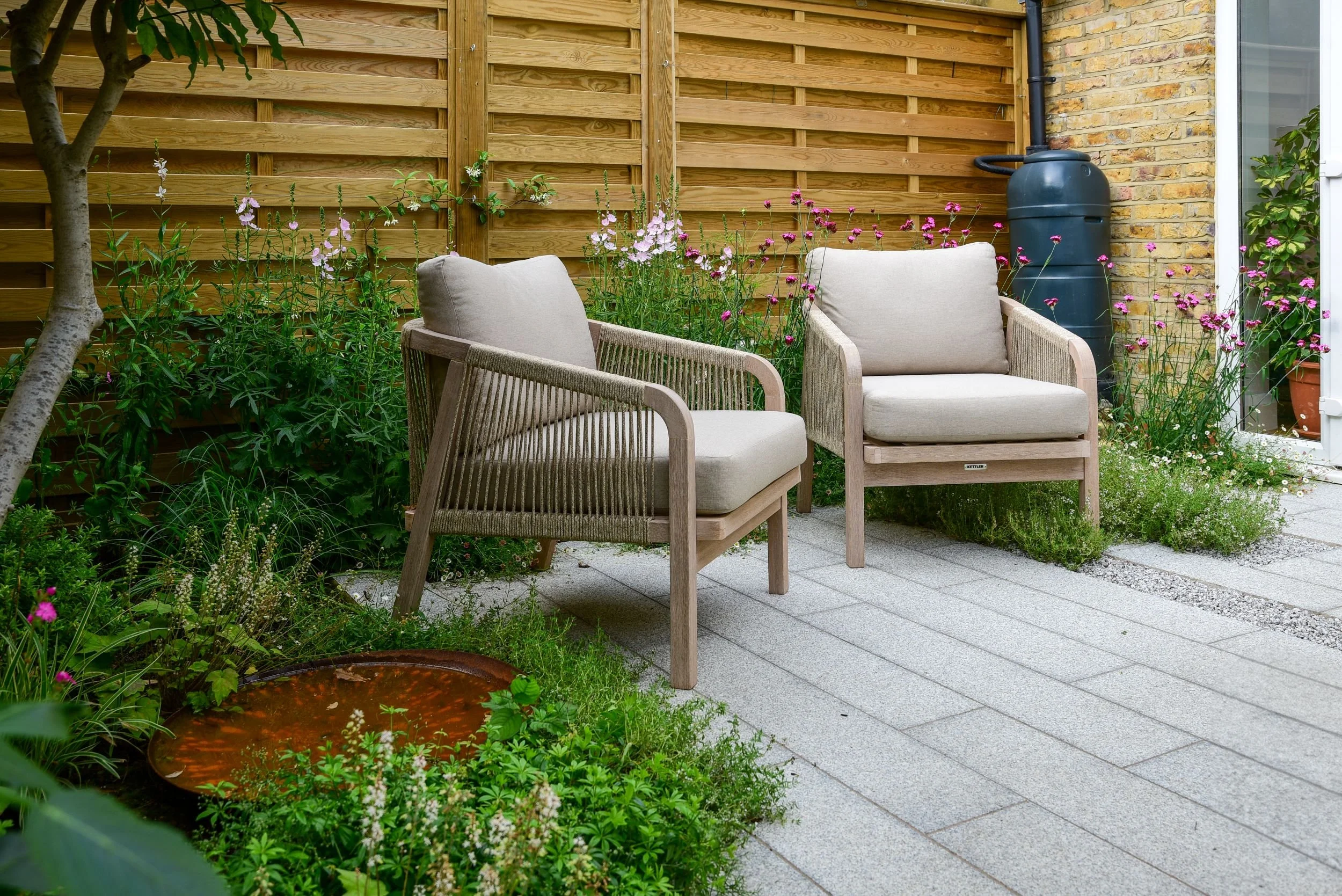 Two lounge chairs beside a corten steel water bowl set into a planted courtyard border

A relaxed seating area pairs neutral rope-weave chairs with a rusted corten steel water bowl, tucked beneath a multi-stem tree and surrounded by naturalistic plan