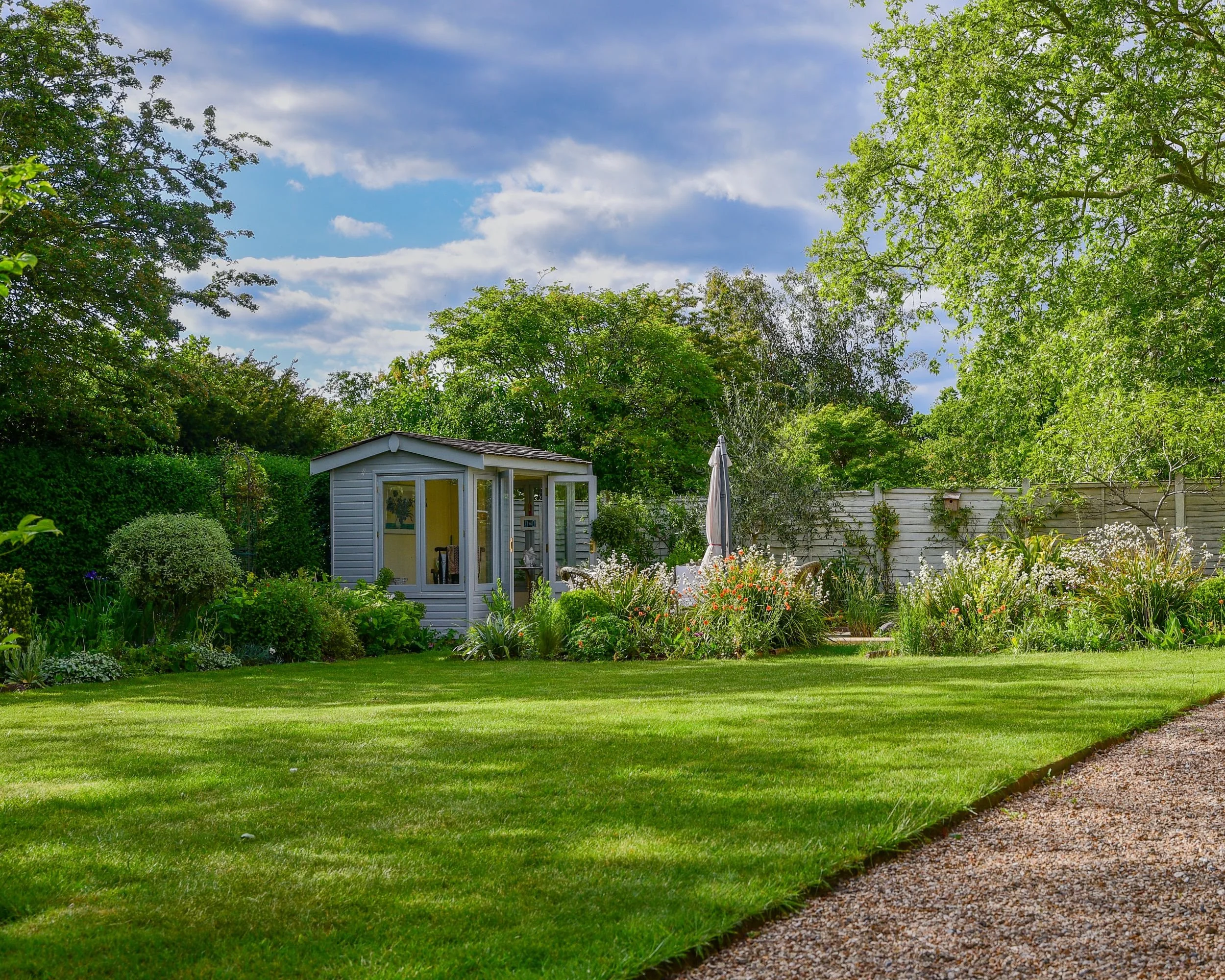 A pale blue-grey summerhouse sits at the edge of a manicured lawn in a Berkshire country garden, framed by mature trees and a curved gravel path. JaneShanksterGardenDesign.co.uk

This wide view captures the garden at its most relaxed, the lawn sweepi