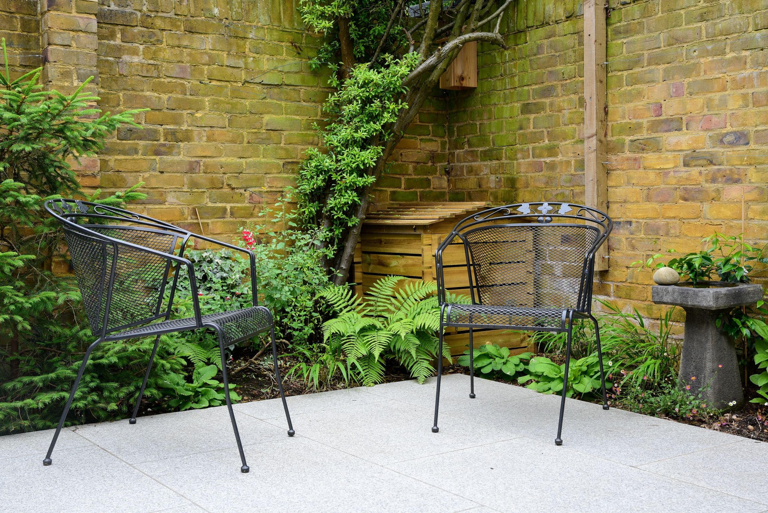 Two black metal garden chairs on granite paving beside ferns and a stone birdbath

A pair of black metal garden chairs sit on honed granite paving in a quiet corner of the garden, surrounded by ferns, roses and a stone birdbath tucked against the rec