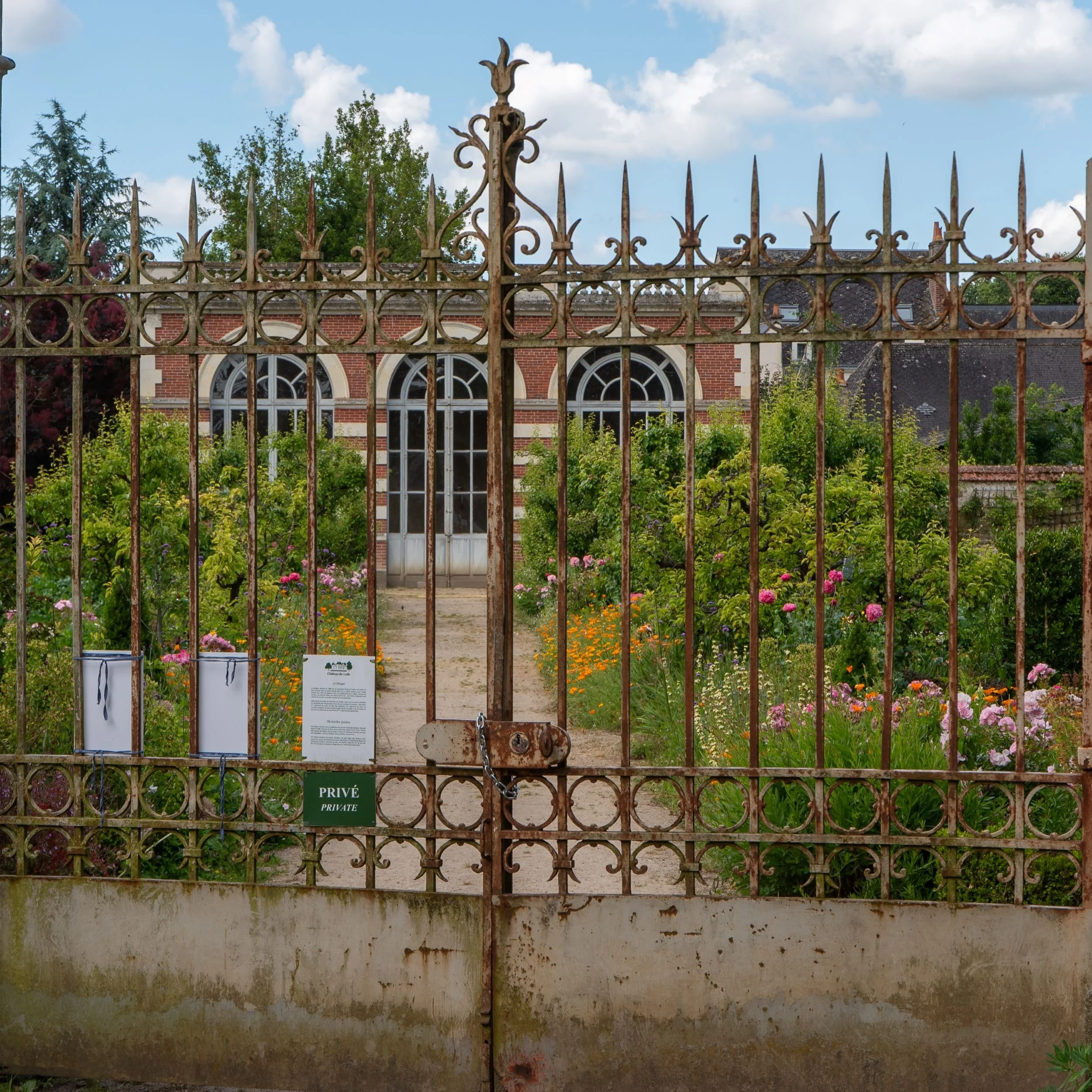 Ornate wrought iron gate marked 'Privé / Private' at Château du Lude, looking through to the kitchen garden and brick orangery beyond