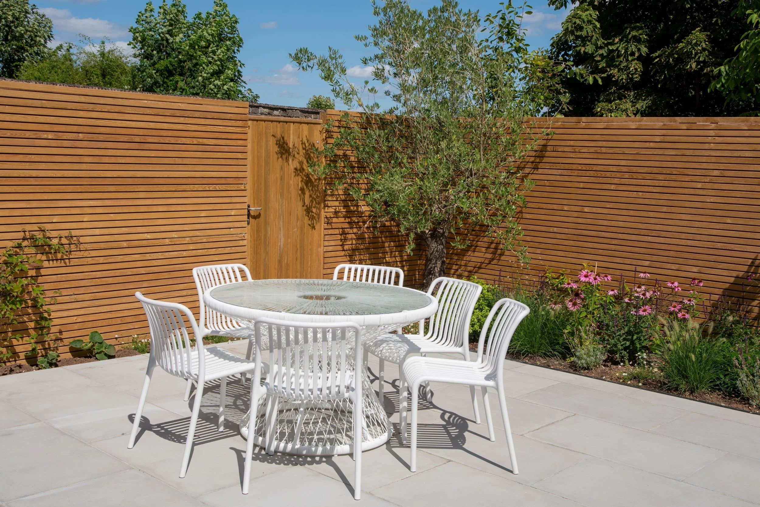 Round dining table and chairs on garden patio, property development

A sunlit dining terrace for a property development garden, centred on a round glass-topped table with white woven chairs. Cedar slatted fencing and a timber gate frame the space, wi