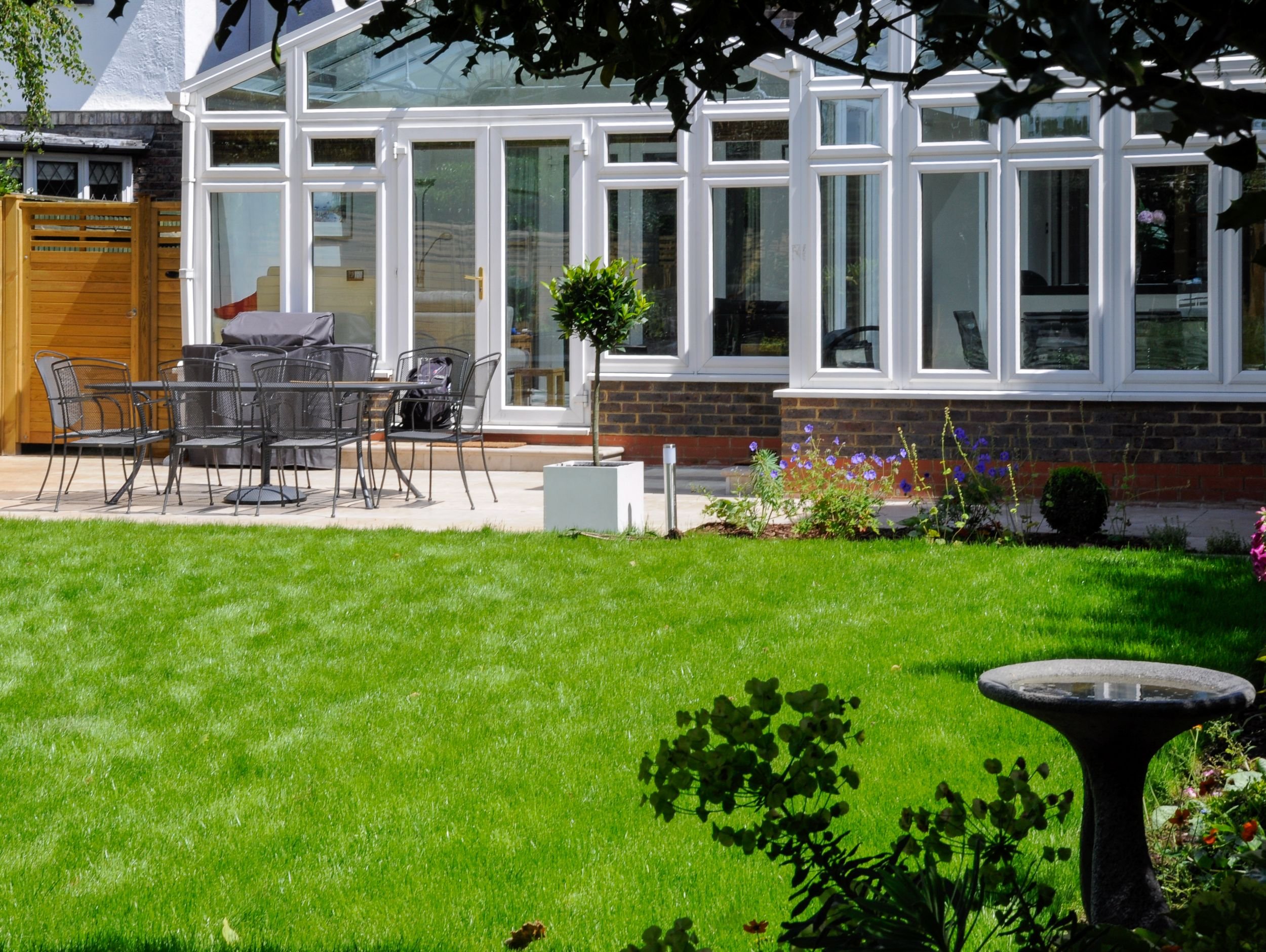 A dining patio with metal table and chairs sits in front of a large conservatory extension in a Raynes Park family garden. JaneShanksterGardenDesign.co.uk

A paved terrace furnished with a metal dining set and a covered barbecue runs along the length