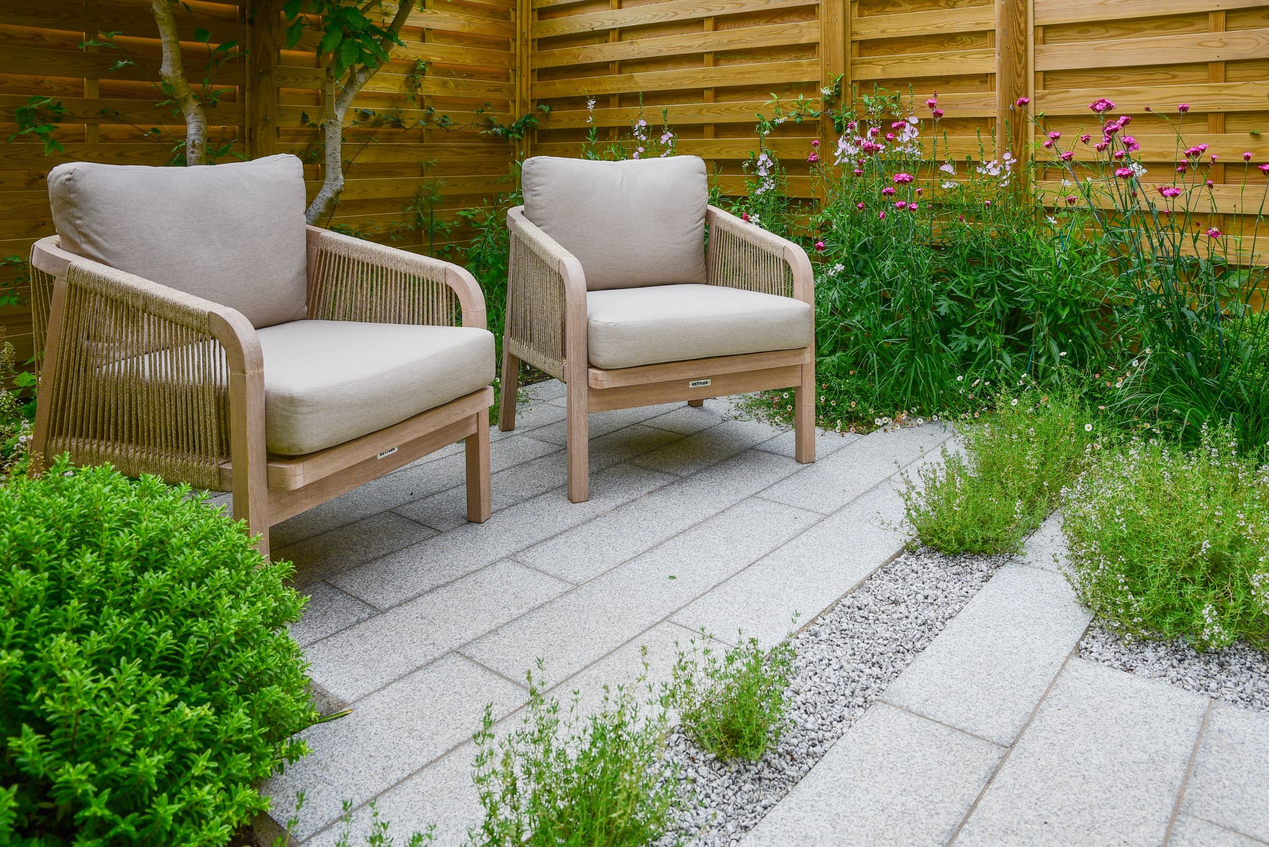 Two woven rope lounge chairs on a granite paved patio surrounded by pink cottage garden flowers

A pair of Kettler rope-weave lounge chairs sit on honed granite paving, framed by timber slatted screening and a colourful border of pink dianthus and si