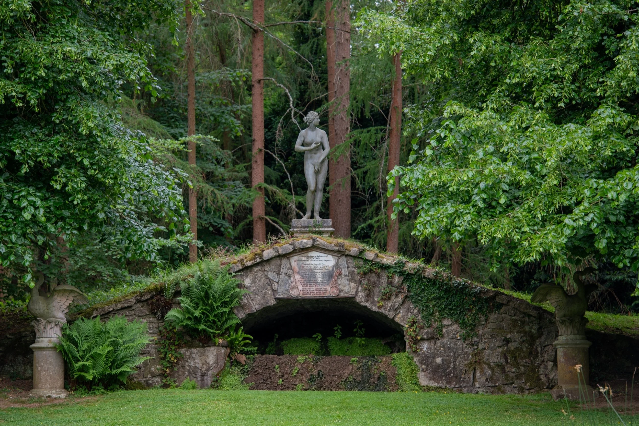 Statue of Venus standing above a moss-covered arched cascade in the Vale of Venus at Rousham, with swan sculptures, ferns and woodland behind.