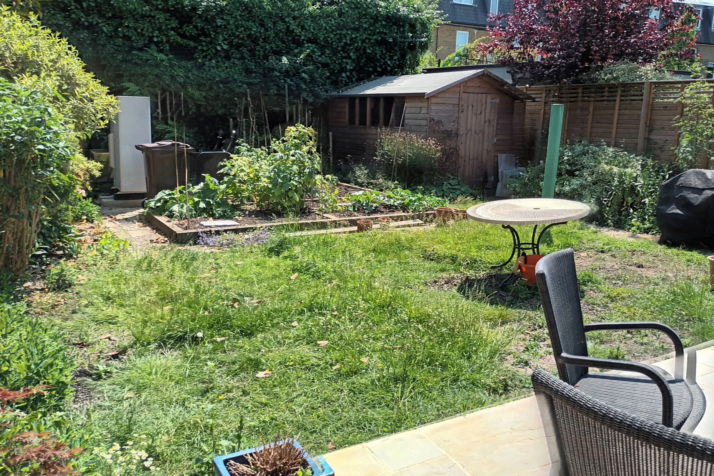 The garden before the redesign: an uneven lawn, an oversized shed, and no defined growing space.

Before photo of an overgrown lawn with a patio table, wheelie bins and a large timber shed at the back of the garden. Designed by JaneShanksterGardenDes