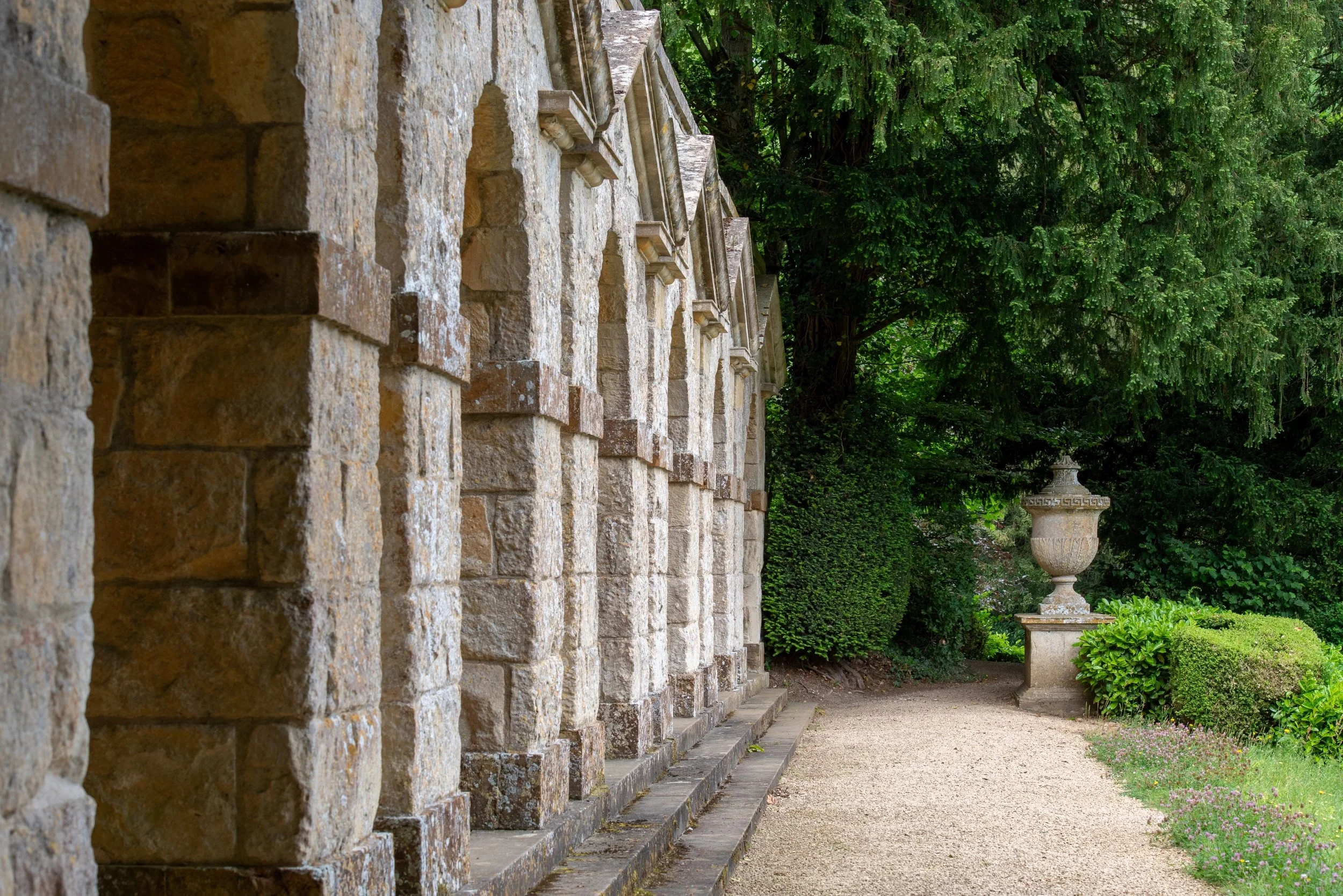 The arcade of the Praeneste Terrace at Rousham, with weathered stone arches beside a gravel path, a clipped yew hedge and a stone urn.