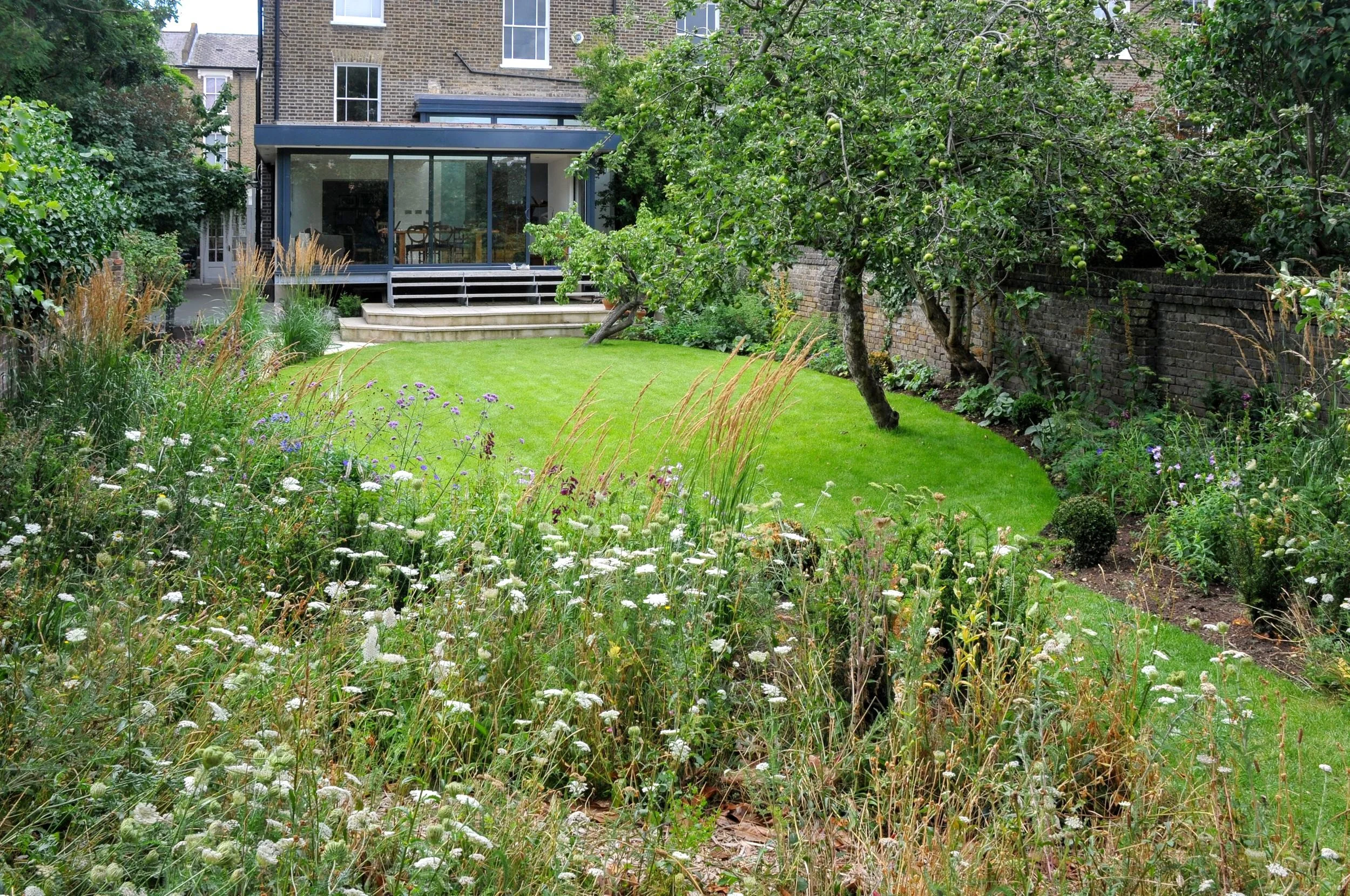 A wildflower meadow of grasses and verbena fills the foreground of a London country garden, with a lawn and house beyond. JaneShanksterGardenDesign.co.uk.

Long grasses and drifts of purple verbena crowd the foreground here, giving way to a neat lawn