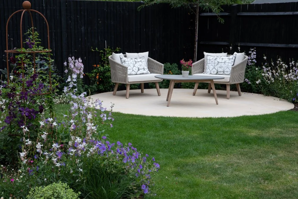 A wide view of a Surrey country garden showing a circular stone terrace with rope-weave chairs, a round coffee table and a clematis-clad rusted obelisk, with lush mixed borders and a neat lawn in the foreground.

 ArthurRoadLandscapes.co.uk. This wid