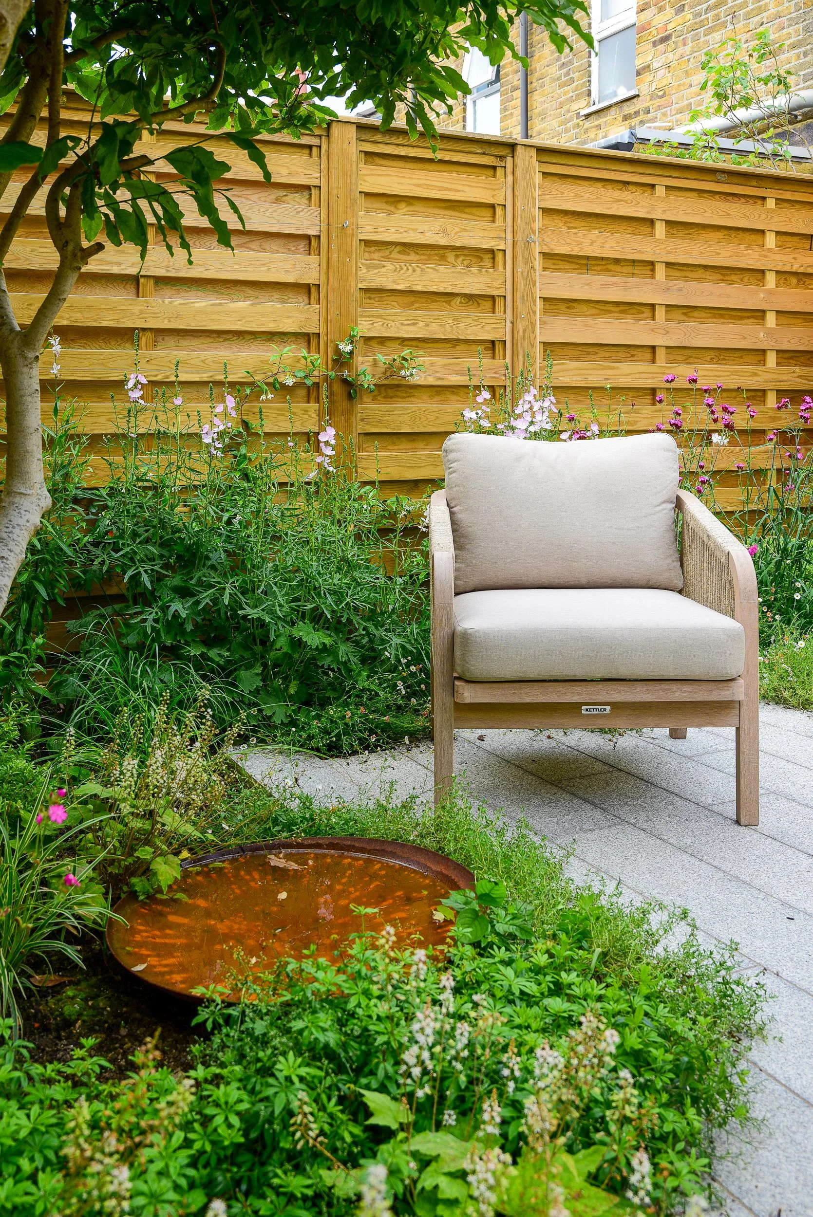 A lounge chair on granite paving beside a corten steel water bowl surrounded by planting

A single Kettler lounge chair overlooks a corten steel water bowl set within a lush planted border, illustrating how seating and water feature combine in this s