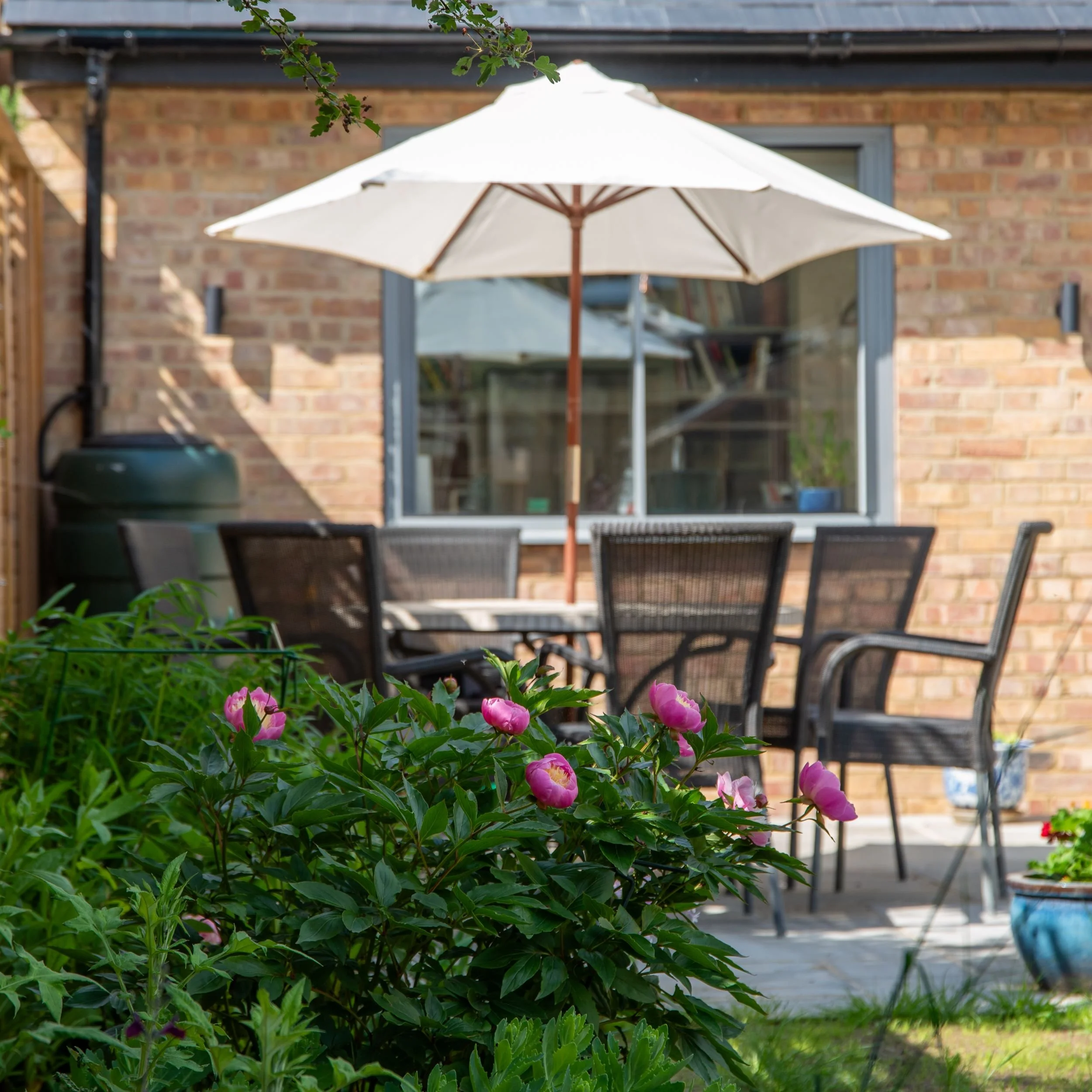 Patio dining table with parasol and chairs against the house, with pink peony flowers in the foreground.

The seating area by the house, with peonies in flower and the extended paving providing space for the dining table. Designed by JaneShanksterGar