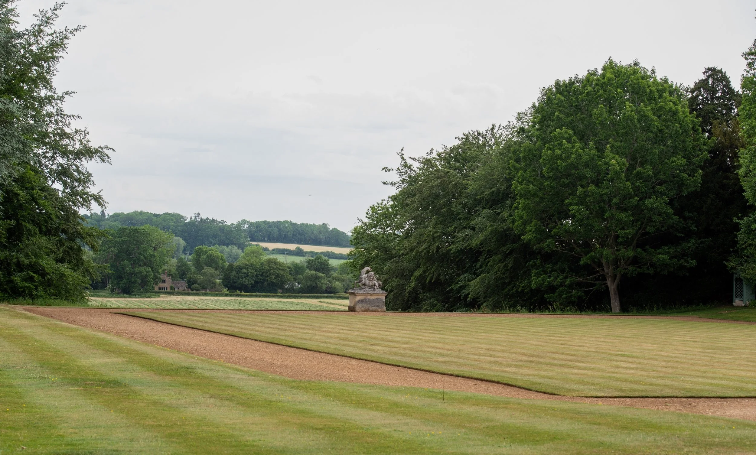 Striped lawn and gravel path of the bowling green at Rousham, leading to a statue of a lion attacking a horse, with Oxfordshire countryside beyond.