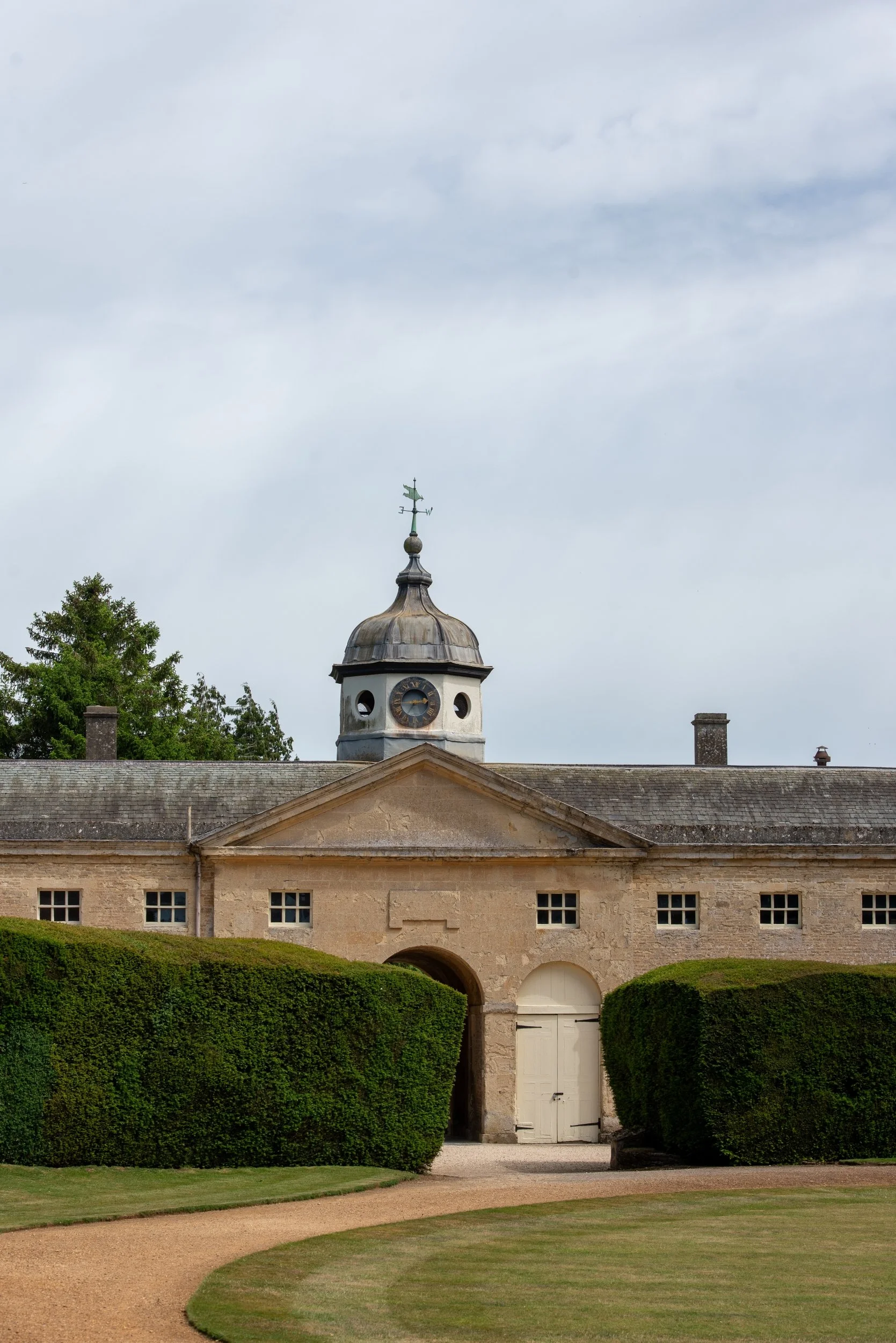Stone stable block at Rousham with a pediment, arched entrance and clock cupola, framed by two clipped yew hedges.