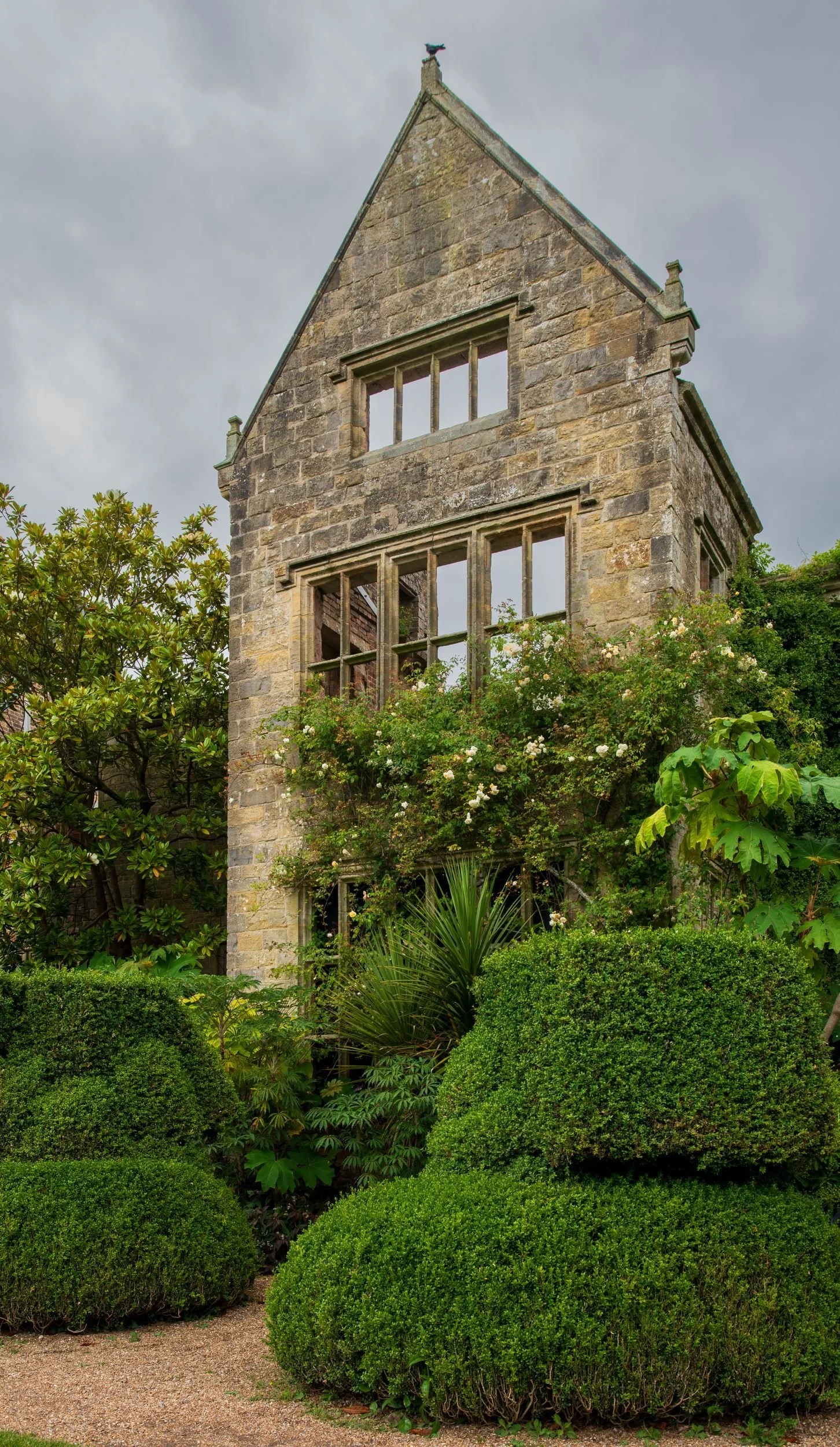 Ruined gable end of Nymans house with empty window frames, climbing roses and clipped box hedges in the foreground.