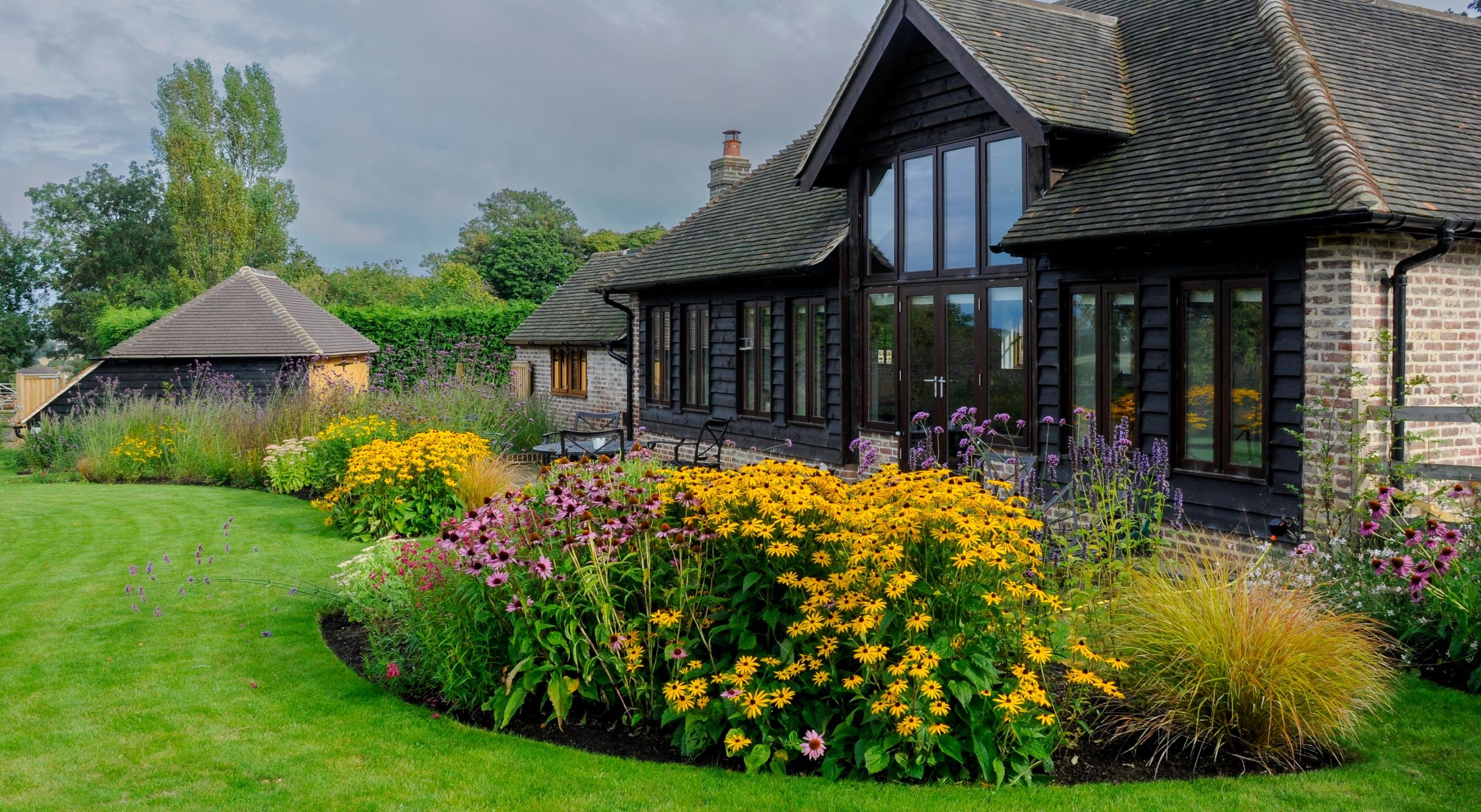 A curving border of rudbeckia, echinacea and ornamental grasses wraps around an oak-framed house in a modern Surrey country garden. JaneShanksterGardenDesign.co.uk.

The same house, transformed: a sweeping curved border of golden rudbeckia and pink e