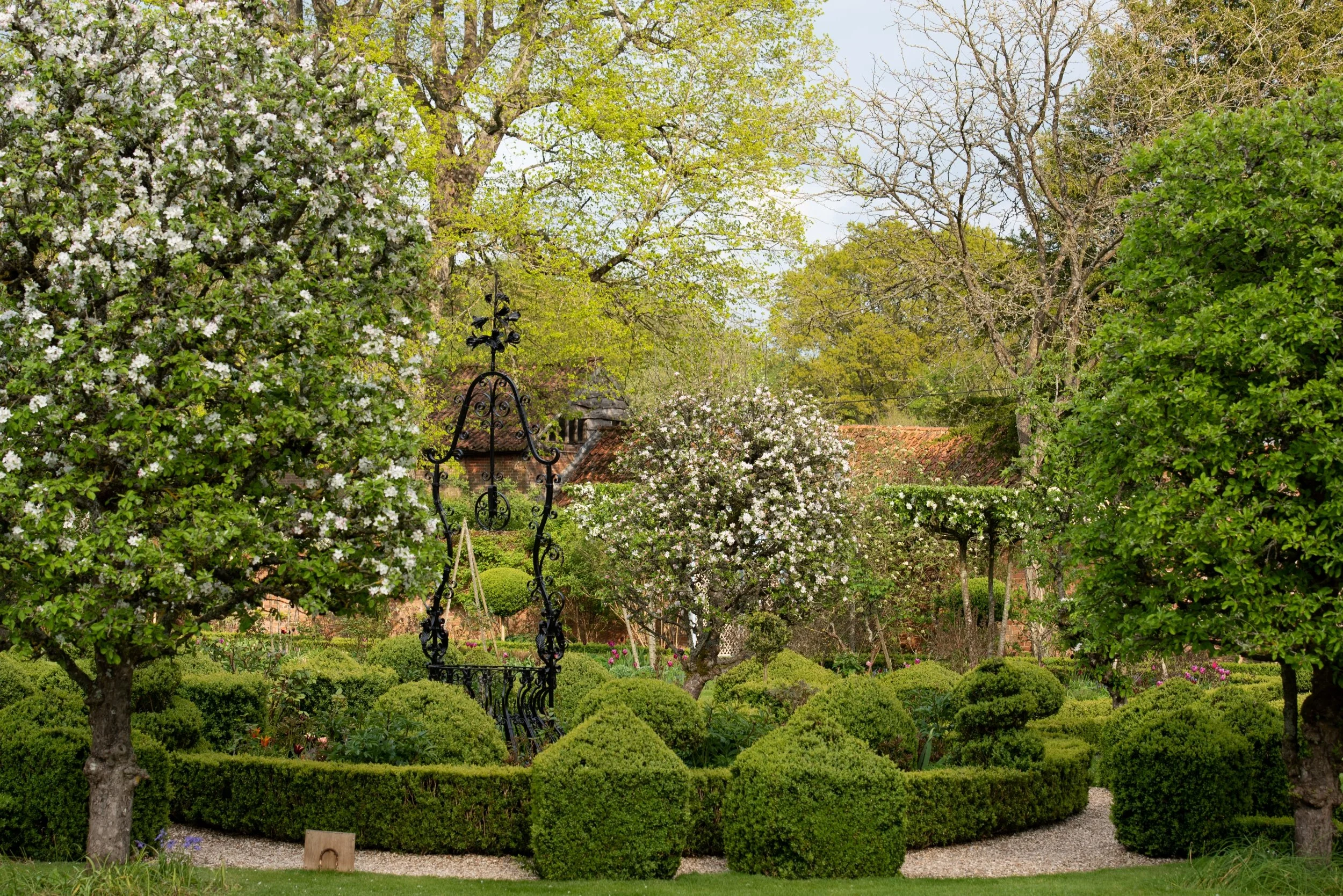 An ornate black metal obelisk rises above clipped box topiary at West Green House Garden, framed by blossoming apple trees.