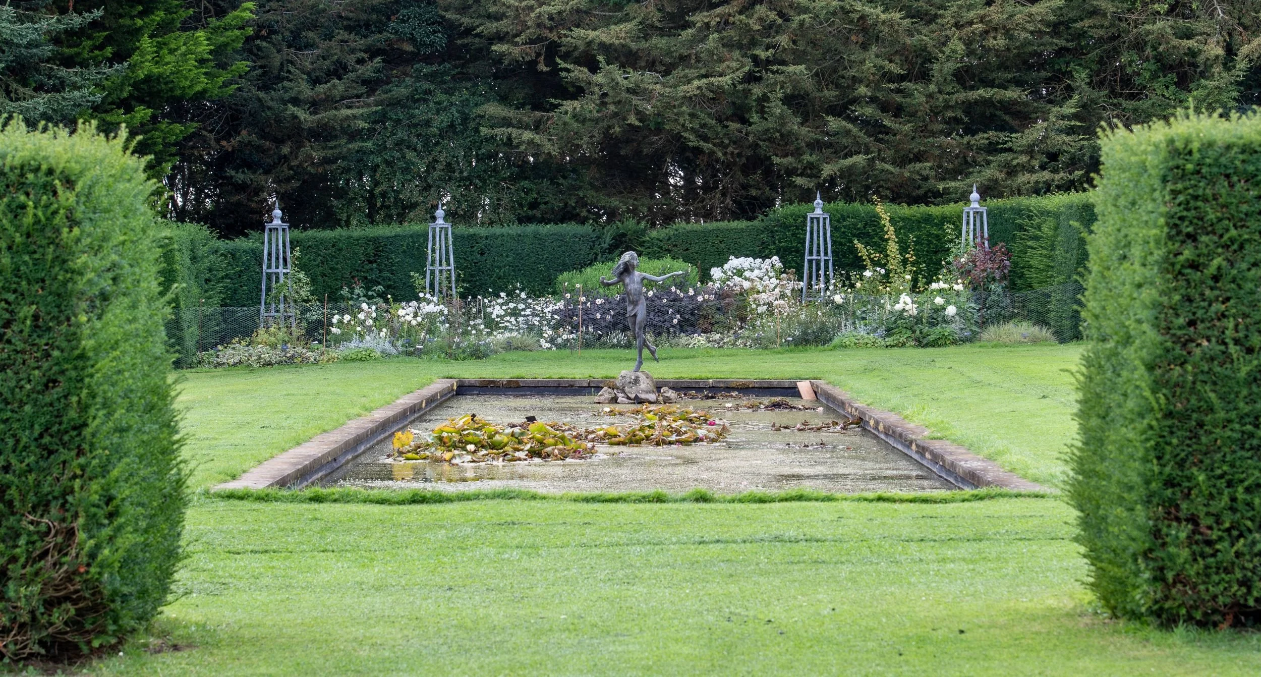 A canal pool at Waterperry Gardens with a bronze figure of a running girl, water lilies, and white and dark herbaceous borders behind.