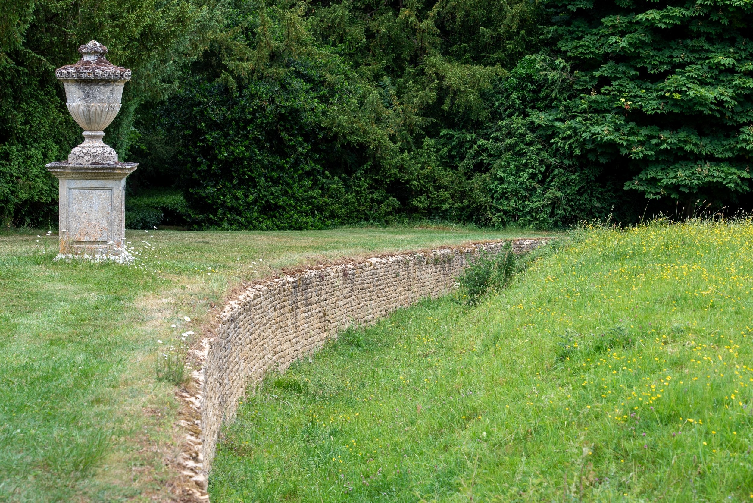 Curving dry stone ha-ha wall at Rousham, with a stone urn on a pedestal and a buttercup meadow on the far side.