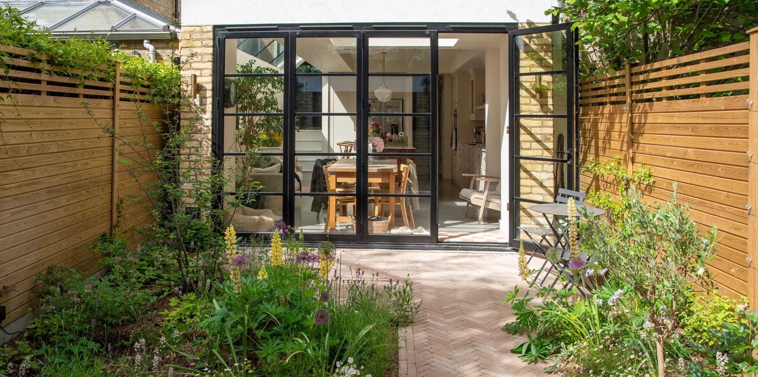 A full view of a Fulham courtyard garden with Crittall-style doors open to the kitchen, herringbone brick paving, naturalistic planting borders of lupins and alliums on both sides, cedar slatted fencing and a bistro table and chairs. ArthurRoadLandsc