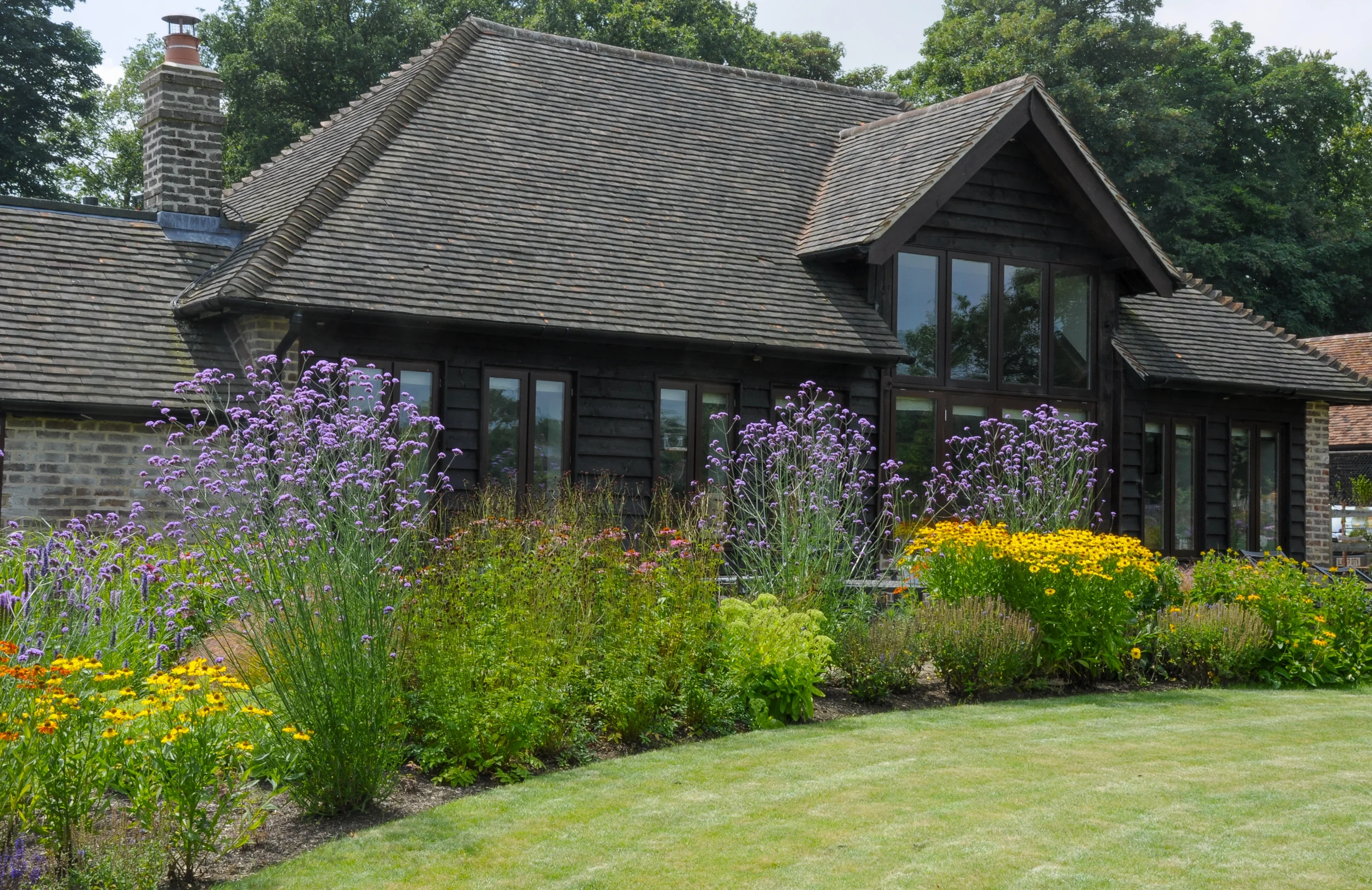 A house with a shingled roof, black exterior wood siding, and large windows, surrounded by a colorful flower garden and a well-maintained lawn, with green trees in the background.