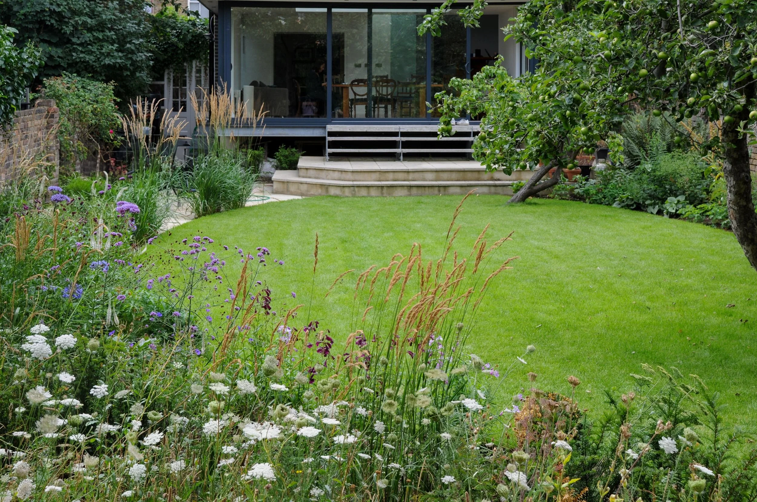 A view across a wildflower border and lawn towards a glass-fronted extension in a London country garden. JaneShanksterGardenDesign.co.uk.

From ground level among the wildflowers, this view takes in the full sweep of the garden back towards the house