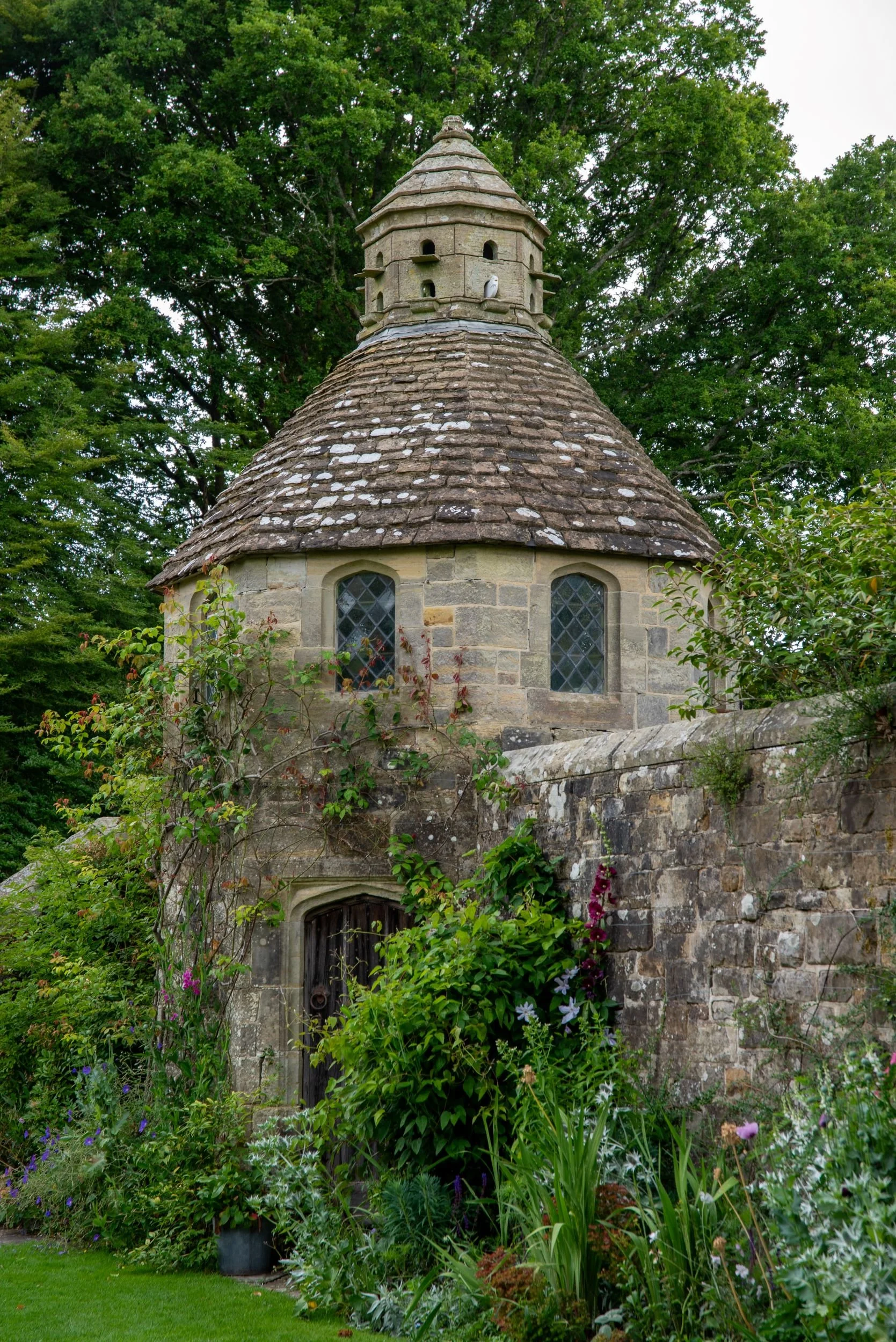 Stone dovecote with a conical tiled roof and lantern nesting tower rising above the garden wall at Nymans, with climbing roses and hollyhocks below.