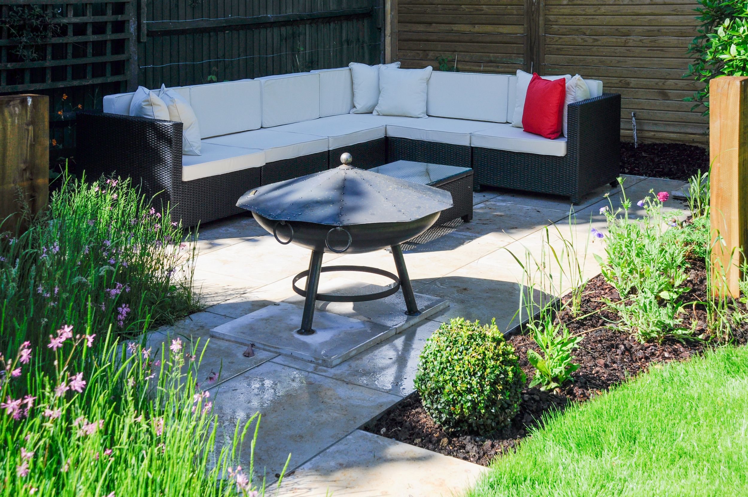 A black rattan corner sofa with cream cushions surrounds a domed firepit on a garden patio. JaneShanksterGardenDesign.co.uk

A generous rattan corner sofa dressed with cream cushions and a bold red accent wraps around a riveted steel firepit, with ca