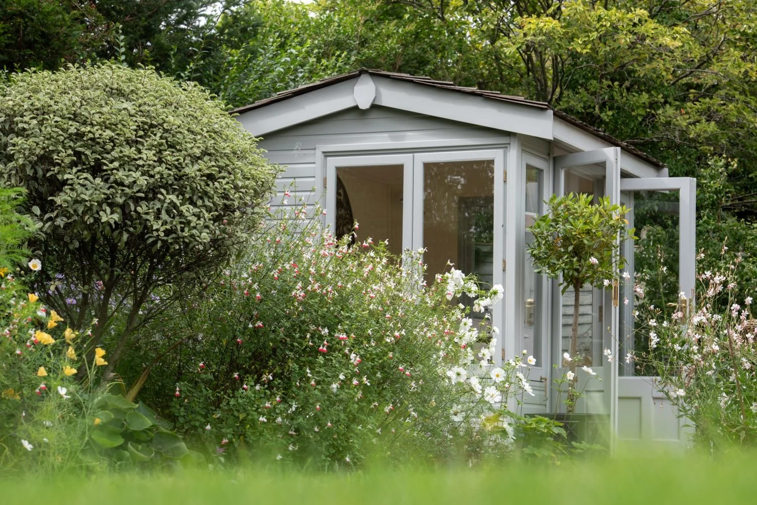A pale grey garden summerhouse with open doors, surrounded by salvia, cosmos and clipped topiary in a Berkshire country garden. JaneShanksterGardenDesign.co.uk.

Seen up close, the summerhouse becomes part of the planting rather than a backdrop to it