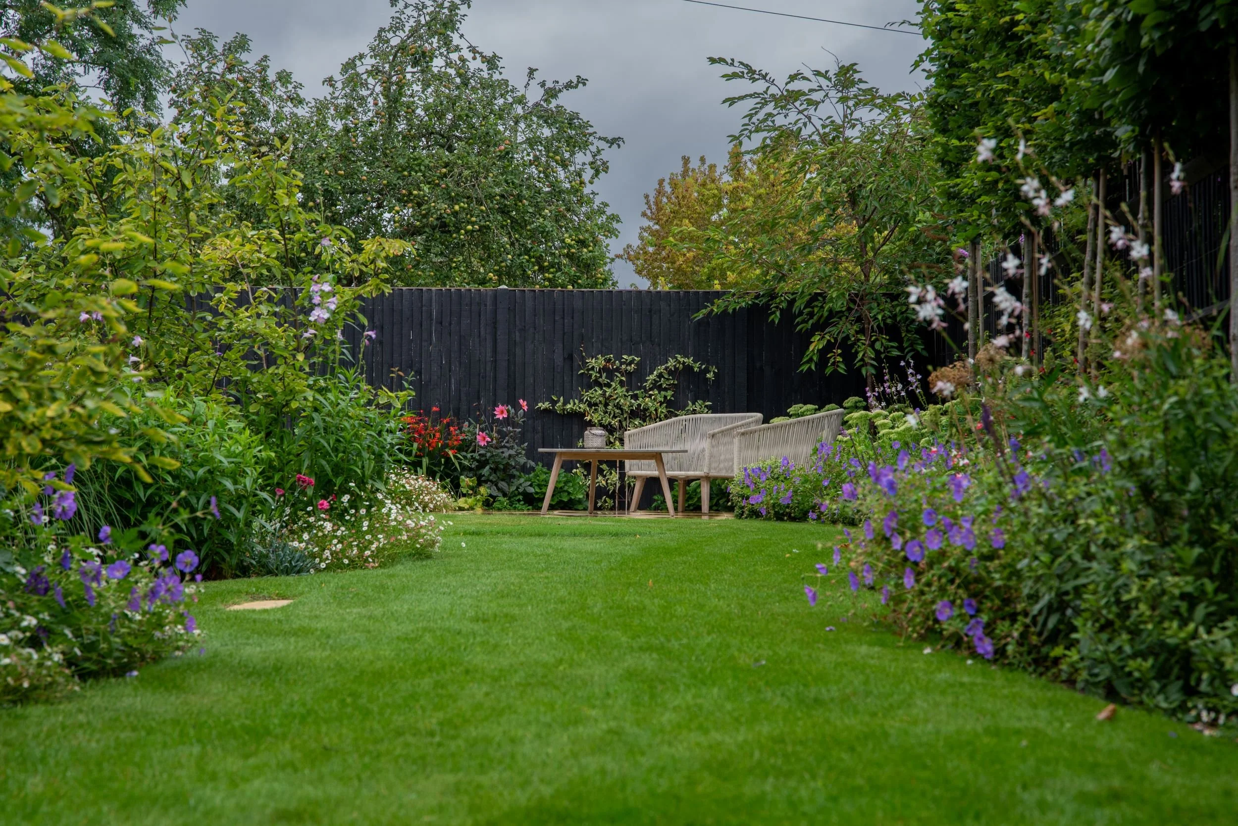 A wide view down a lush green lawn in a Surrey country garden, framed by abundant mixed borders of geranium, cosmos, dahlias and clematis, with rope-weave garden furniture and a dining table set against the black timber boundary fence at the far end.