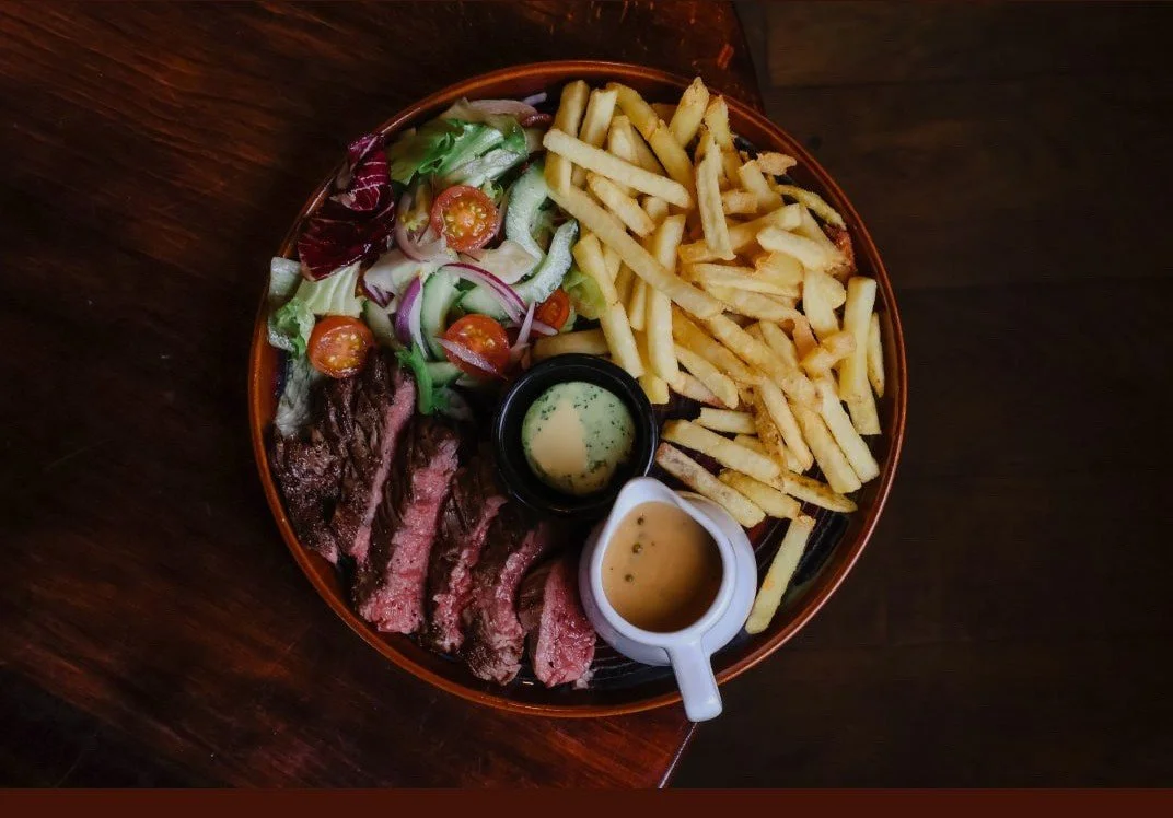 A plate of sliced steak, french fries, mixed salad with cherry tomatoes, and two small containers of sauce, garnished with leafy greens, on a dark wooden table.