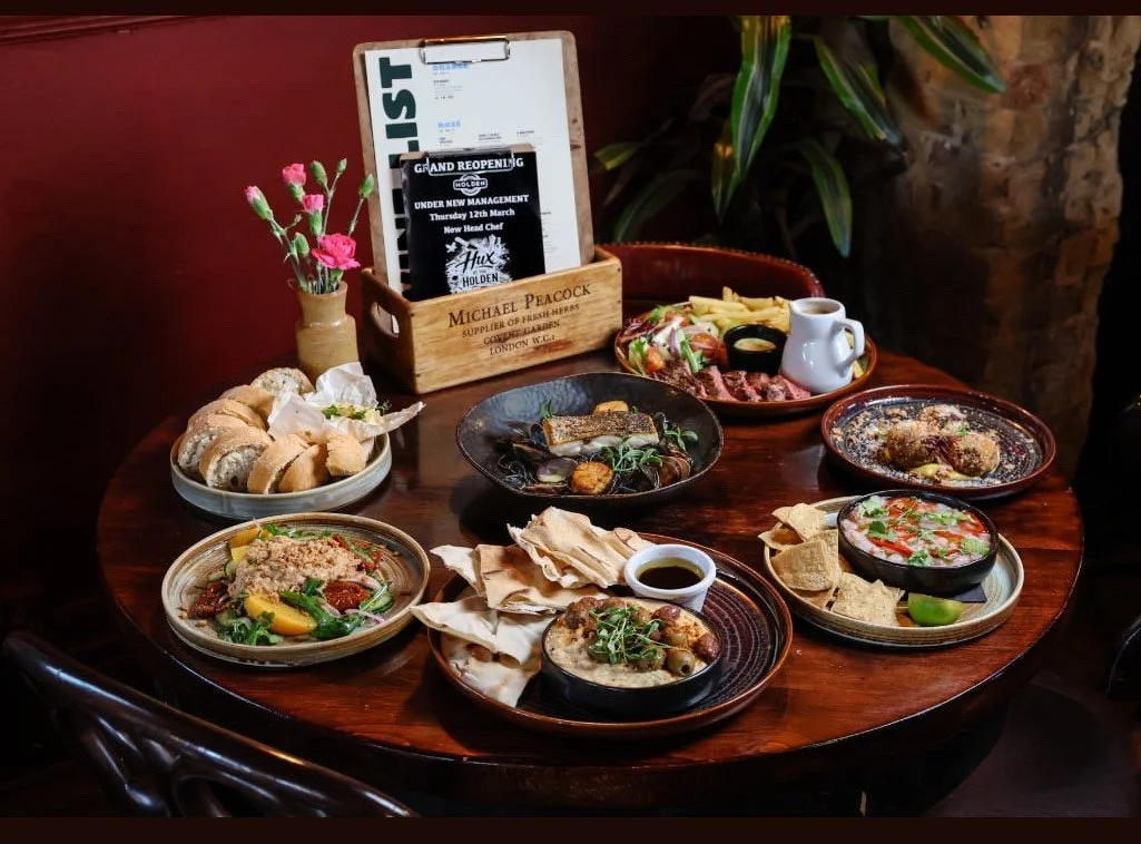 A round wooden table with various plates of food, including bread rolls, a salad, a plate of pasta, grilled meats with vegetables, a bowl of dip with pita chips, and a bowl of soup. There is also a small vase with pink carnations and a wooden box holding a menu or flyer, along with some greenery in the background.