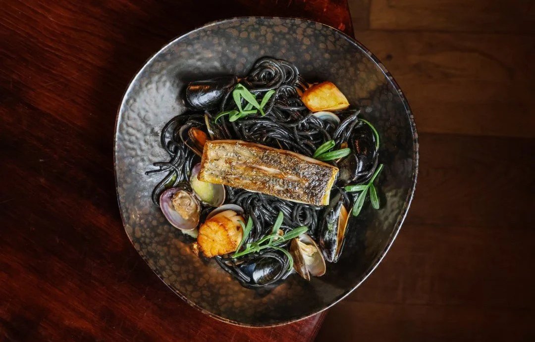 Black squid ink pasta with seafood, garnished with green herbs, served on a black textured bowl.