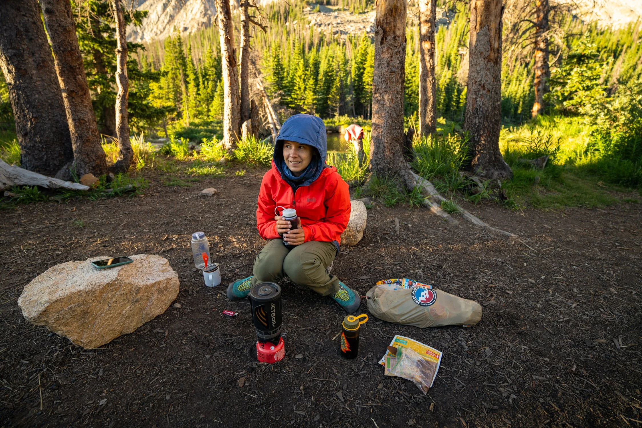 A woman in a red jacket and green pants is sitting on a rock in a forested area with tall trees and green foliage behind her. She is holding a water bottle and smiling. Around her are camping and hiking supplies, including a large bag, a flashlight, 