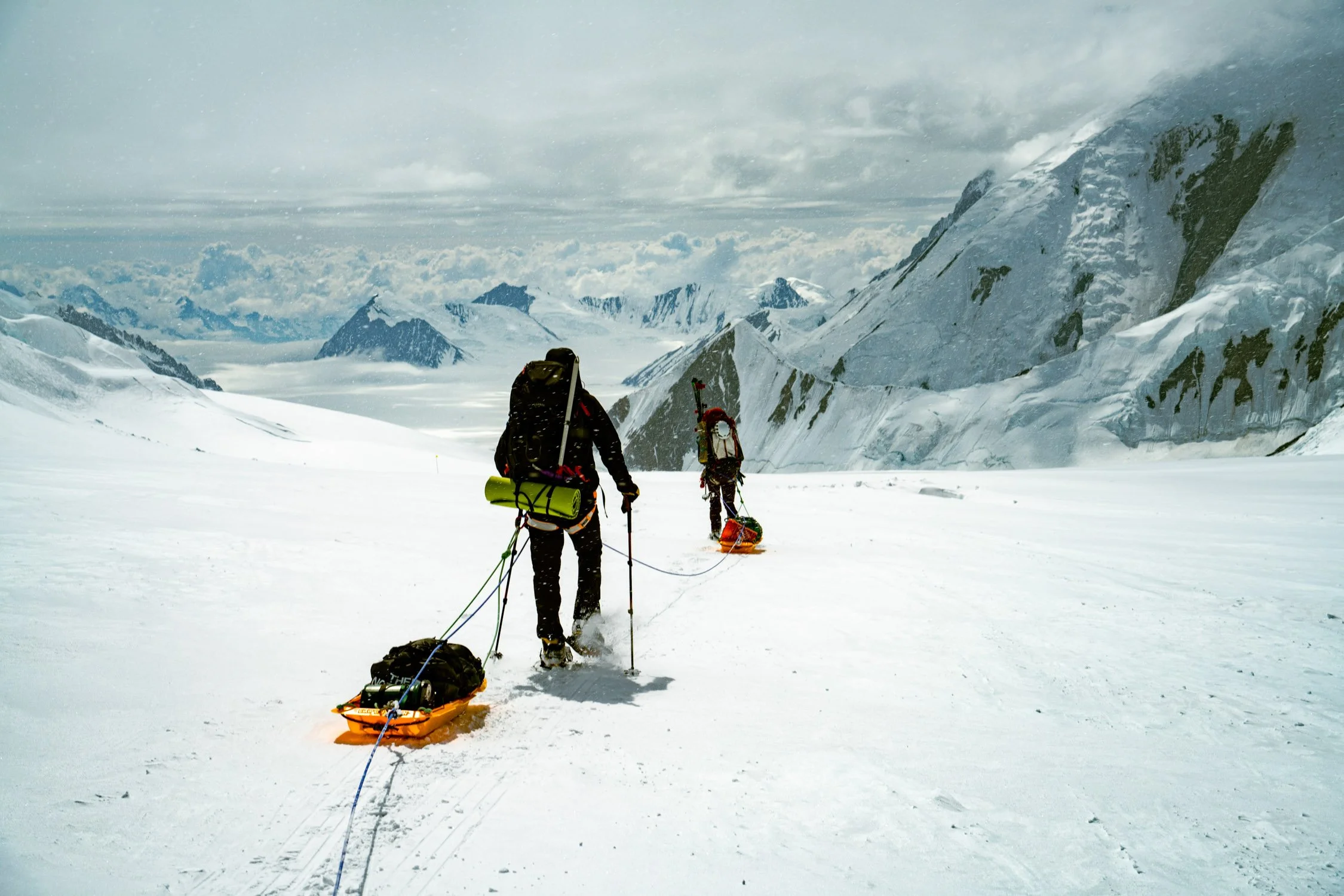Two climbers with backpacks and skis are pulling a rescue sled in a snowy, mountainous landscape with rugged peaks and cloudy skies.