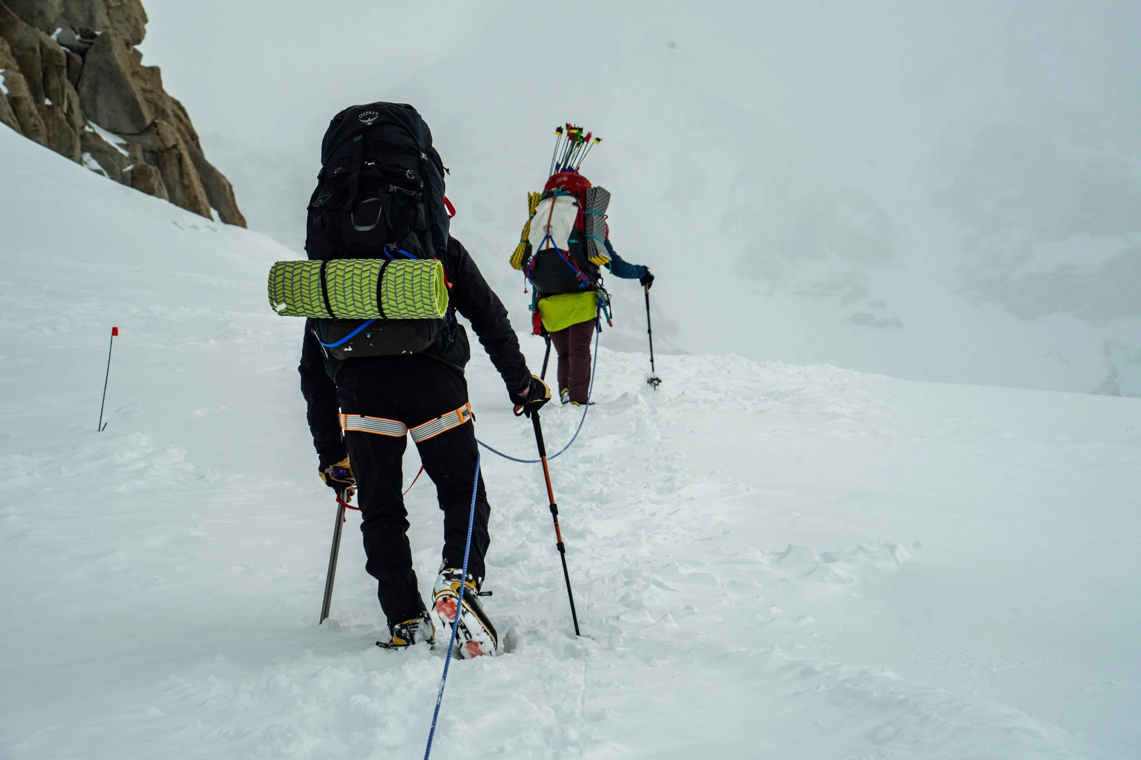 Two mountaineers trekking through a snow-covered mountain with rocky outcroppings, carrying backpacks and hiking poles, heading into foggy weather.
