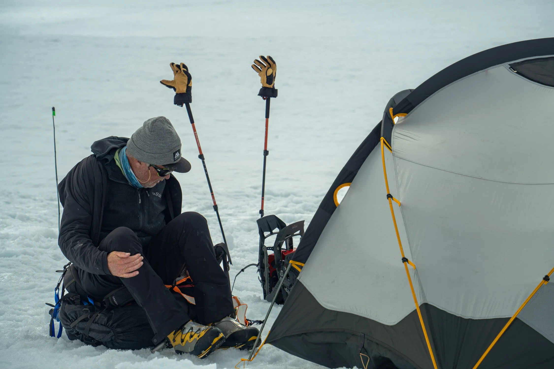 A person dressed in winter clothing is kneeling on snowy ground next to a tent and adjusting his gear. Two trekking poles with gloves are planted in the snow nearby. The scene appears to be in a cold, snowy environment.