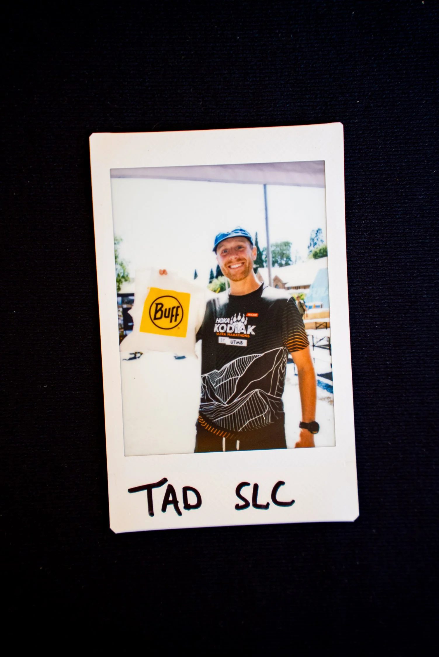 Photograph of a smiling man in a black sports shirt with white and orange patterns, wearing a blue cap and a black watch, standing outdoors in a sunny area with tents and trees in the background, holding a yellow sign with the Buff logo and the text "TAD SLC" written underneath.