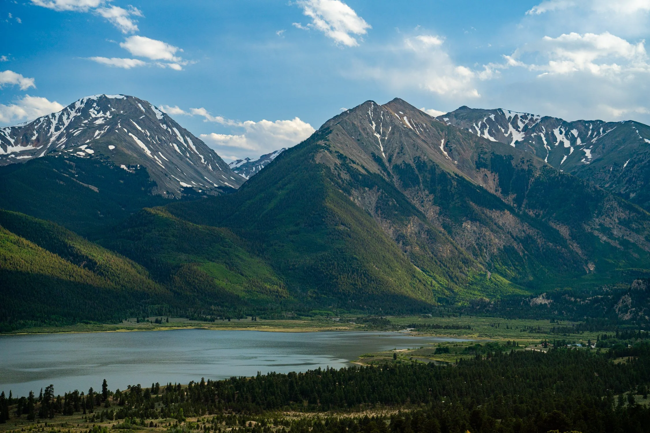 Mountain landscape with snow-capped peaks, green forested slopes, and a large body of water in the foreground under a partly cloudy sky.