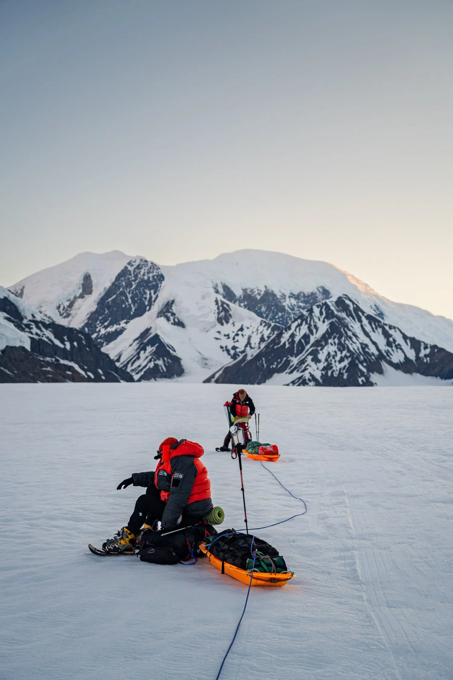 Two mountaineers in red jackets setting up equipment on a snowy glacier with snow-capped mountains in the background during sunset.