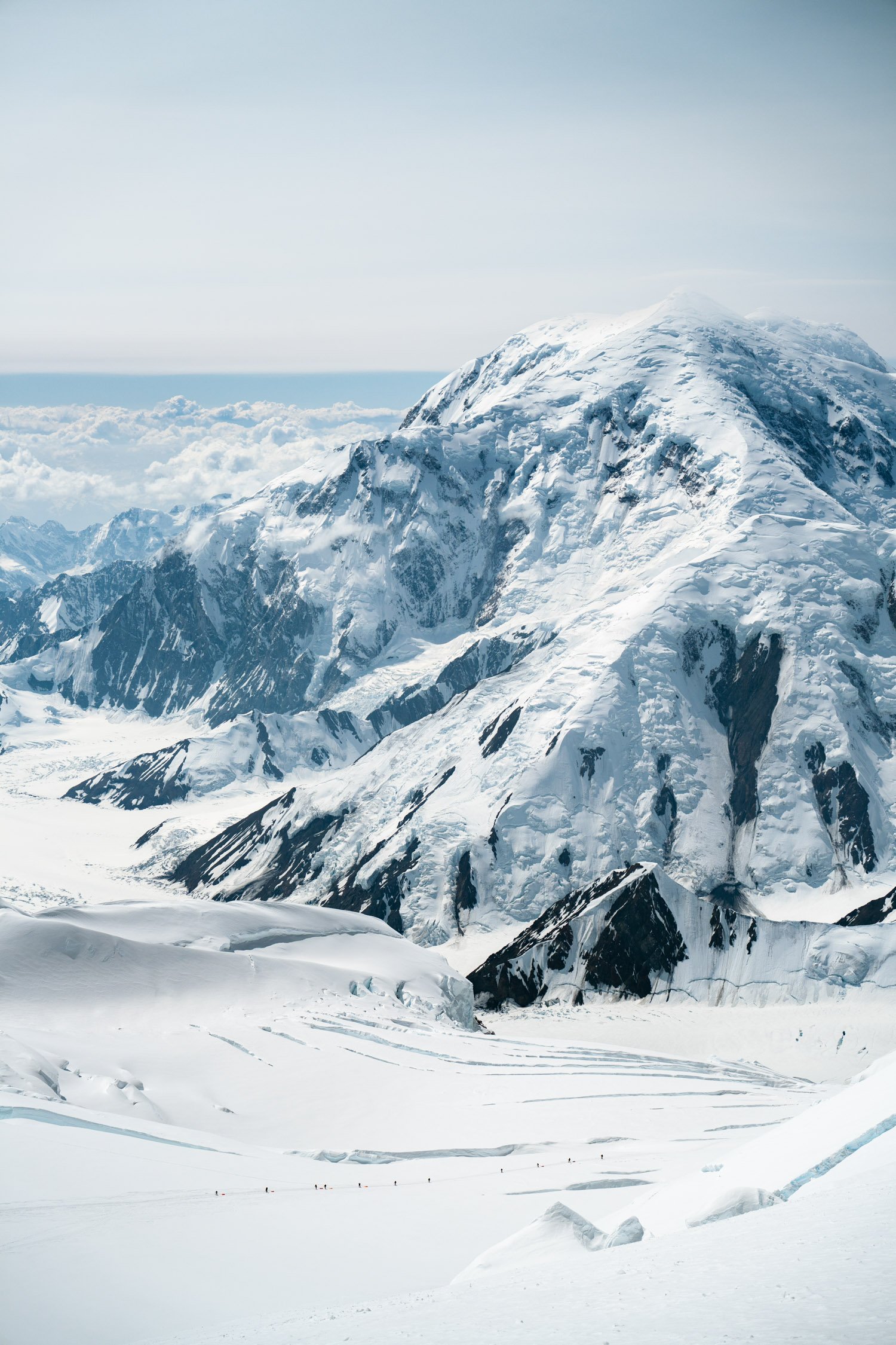 Snow-covered mountain range with steep peaks, glaciers, and a cloudy sky, with a group of tiny climbers or skiers in the distance in the snow.