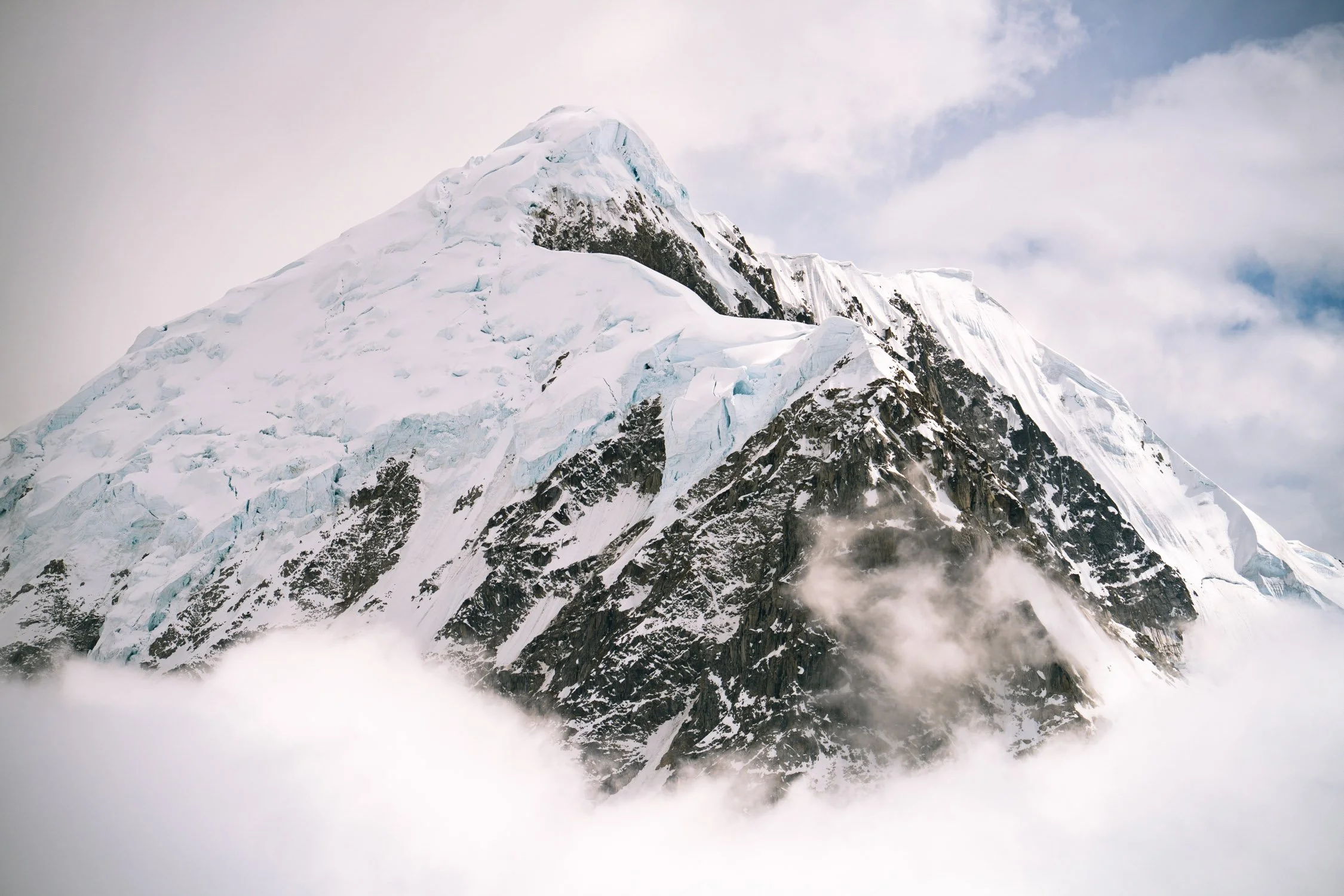 A snow-covered mountain peak with glaciers, rocky slopes, and clouds surrounding the base.
