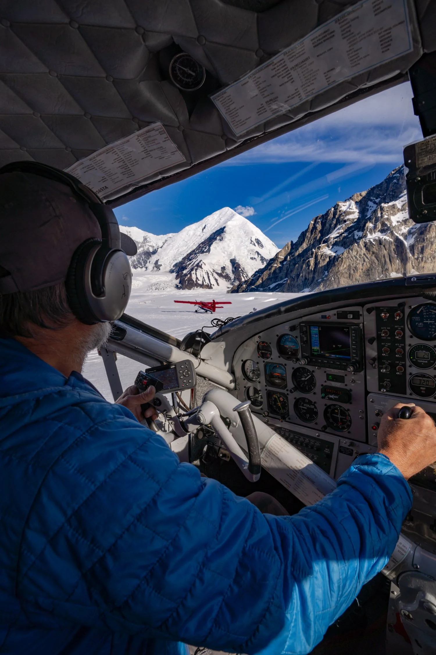 A person wearing a blue quilted jacket and headphones operating a small aircraft, with snow-covered mountains and a red plane on the runway outside the cockpit.