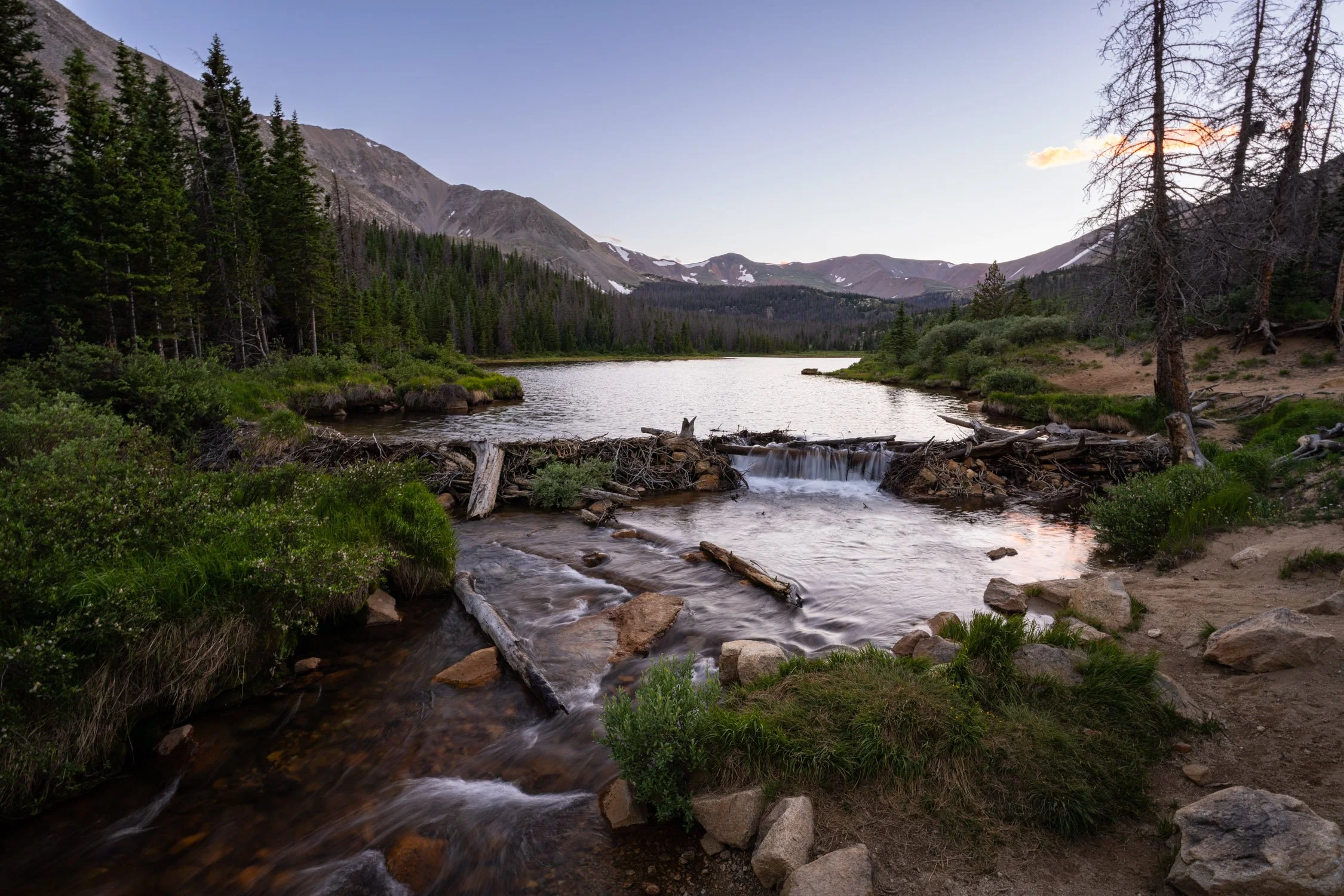 Scenic view of a mountain lake surrounded by evergreen trees, with snow-capped peaks in the background, and a small waterfall flowing over rocks in the foreground.